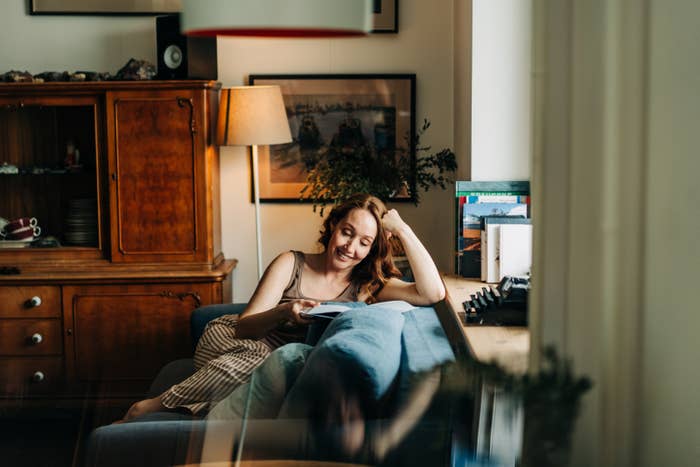 Woman sitting on a sofa in a cozy living room, smiling and reading a book, with a wooden cabinet and framed pictures in the background