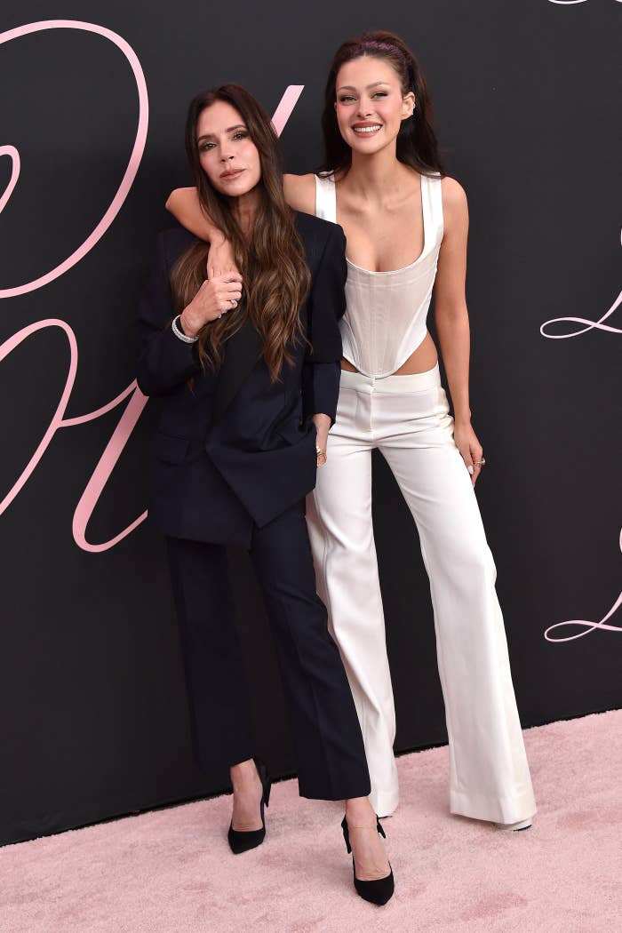 Victoria Beckham dan Nicola Peltz berpose bersama di sebuah acara. Yang kiri mengenakan setelan jas berwarna gelap berstruktur, sedangkan yang kanan mengenakan setelan jas putih dengan potongan