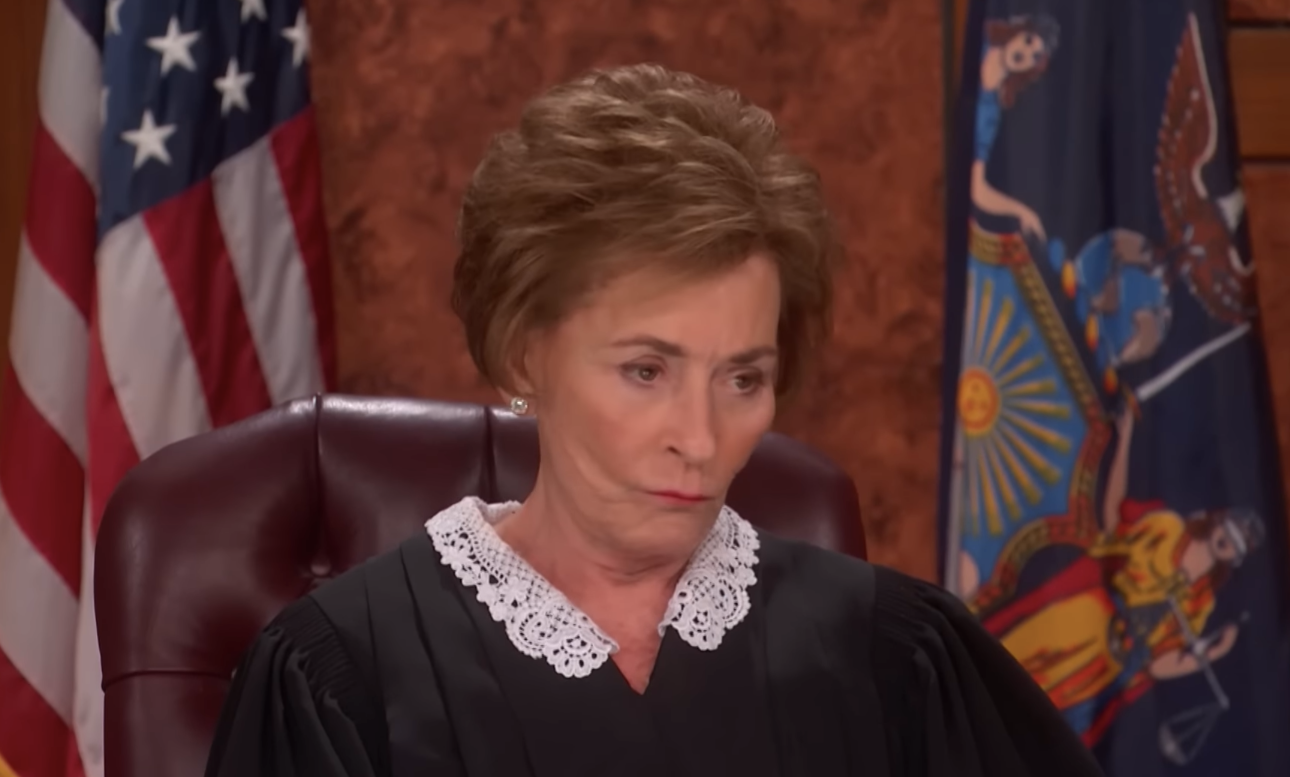 A justice  successful  a courtroom setting, wearing a robe with a lace collar, sits successful  beforehand   of an American emblem  and a authorities   flag