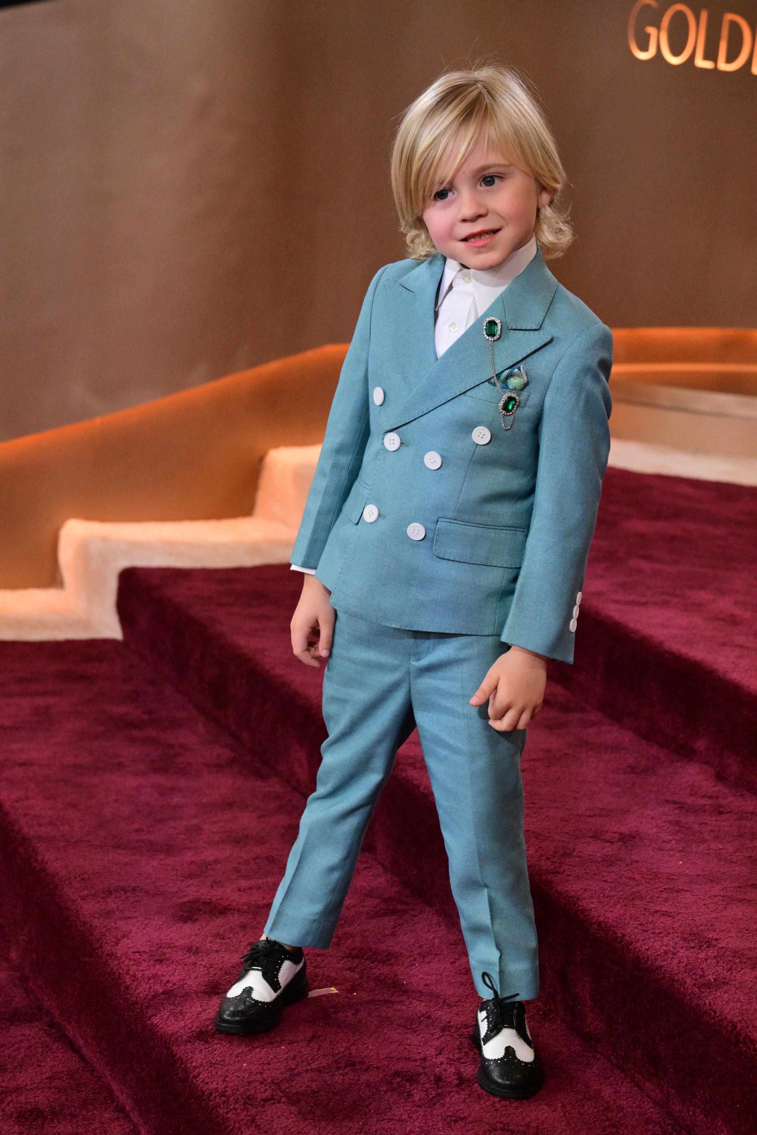 Child on red carpet in stylish suit with brooches, standing confidently and posing for photographers