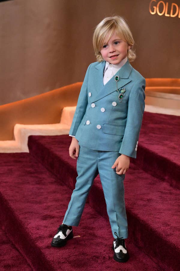 Child on red carpet in stylish suit with brooches, standing confidently and posing for photographers