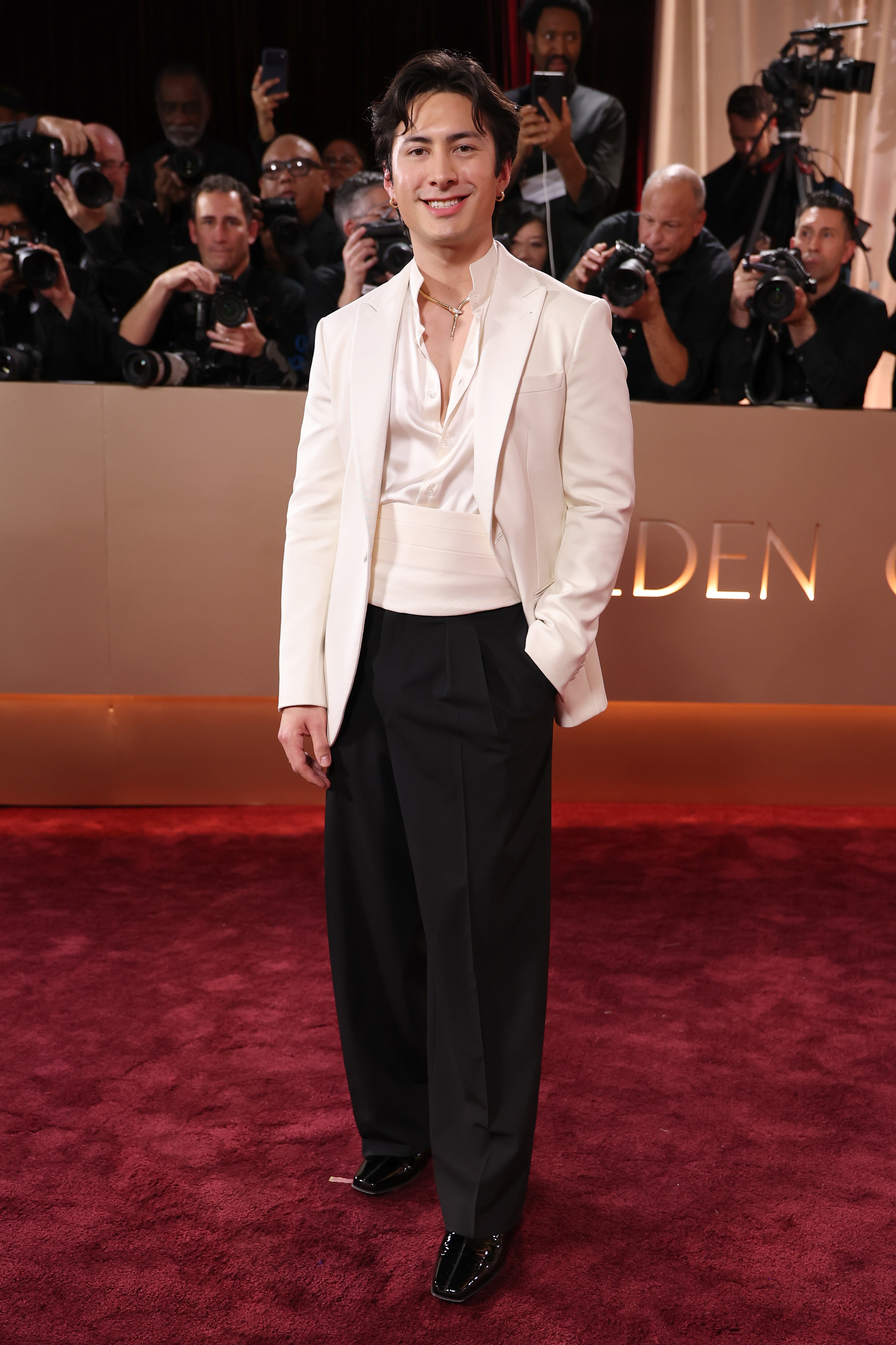 Person in a sleek white tuxedo jacket, black pants, and shiny shoes on a red carpet, surrounded by photographers