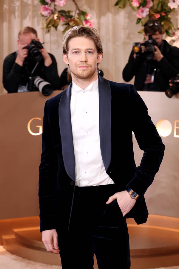 A person on the red carpet in a velvet suit with satin lapels poses for photos, surrounded by photographers