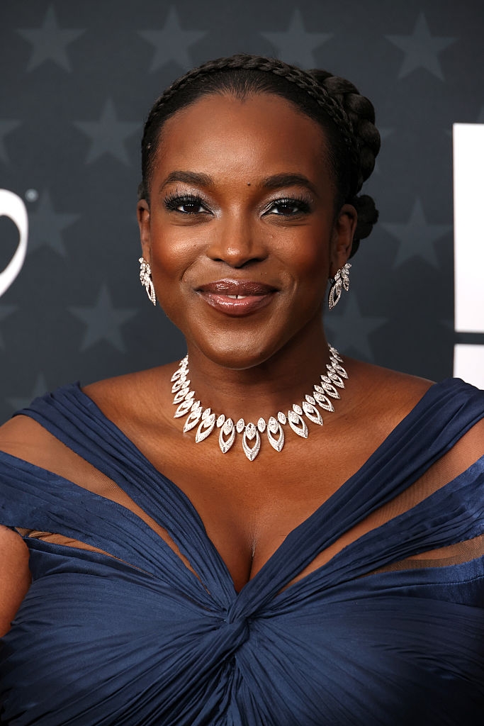 Person smiling connected  a reddish  carpet wearing an elegant gown and jeweled necklace and earrings