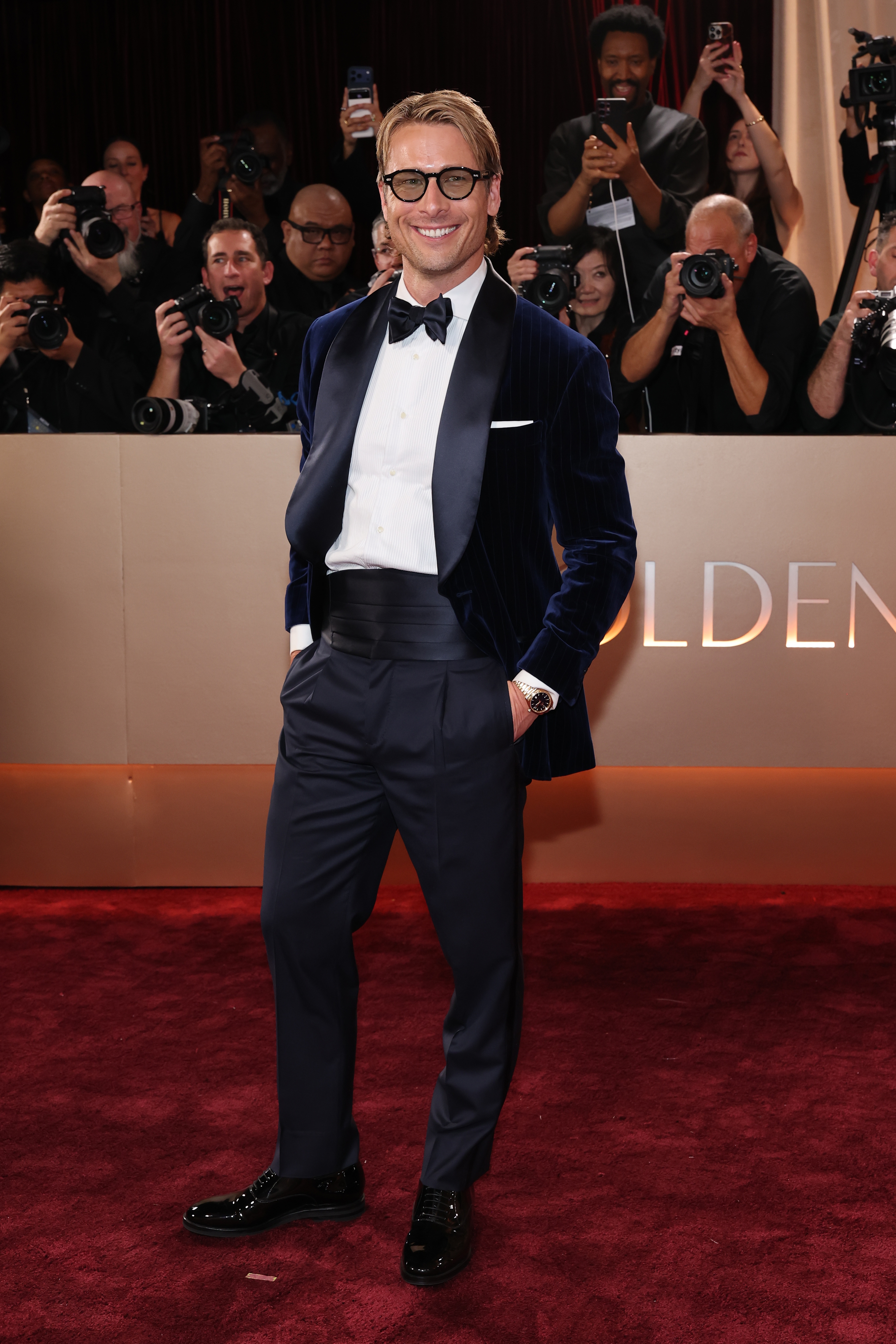 Celebrity on the red carpet in a velvet tuxedo, bow tie, and dress shoes, smiling at photographers.