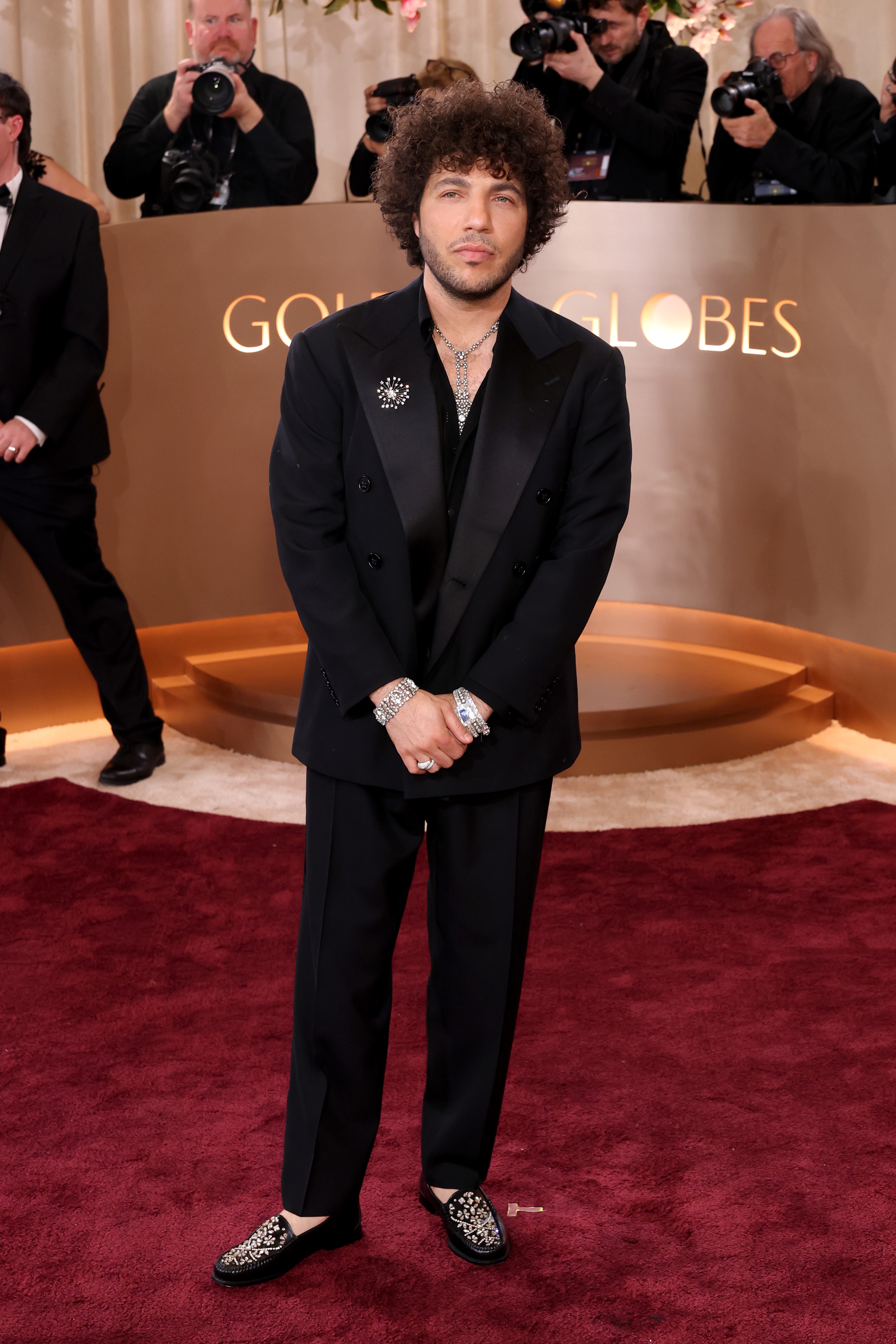 Person in a stylish black suit with sparkling loafers stands on a red carpet, photographers in the background