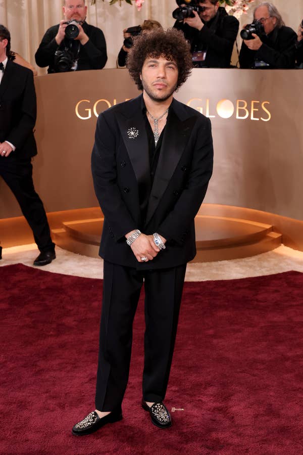 Person in a stylish black suit with sparkling loafers stands on a red carpet, photographers in the background
