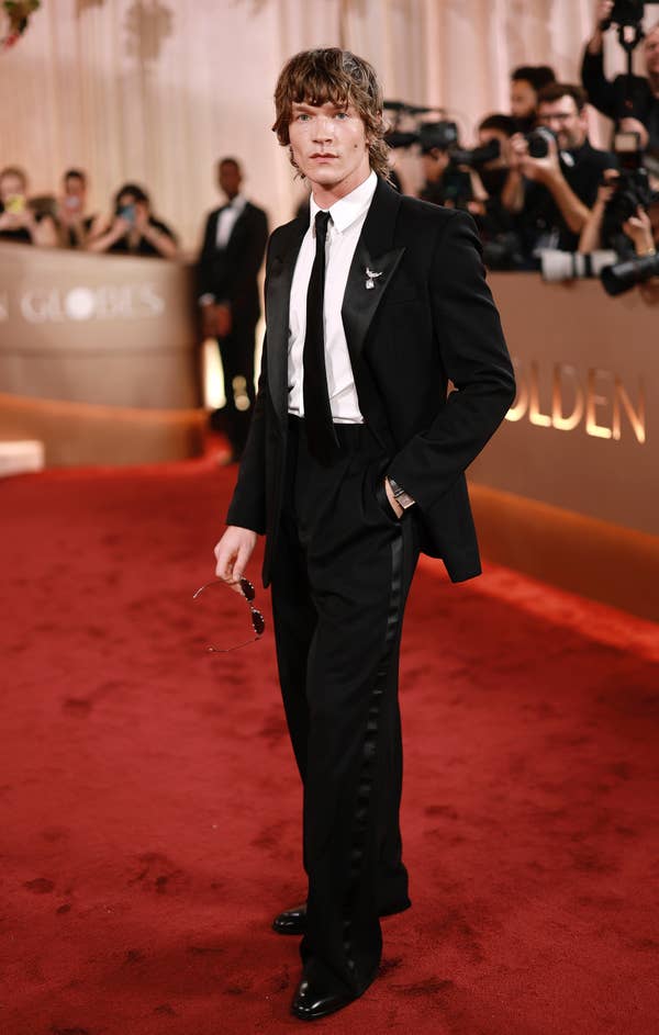 Person on red carpet in classic black suit with white shirt and tie, holding sunglasses. Photographers capture the moment in the background