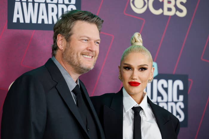 Blake Shelton and Gwen Stefani basal   connected  a reddish  carpet. The pistillate   wears a tailored suit, achromatic  shirt, and necktie  with a bun hairstyle; the antheral   wears a ceremonial  suit
