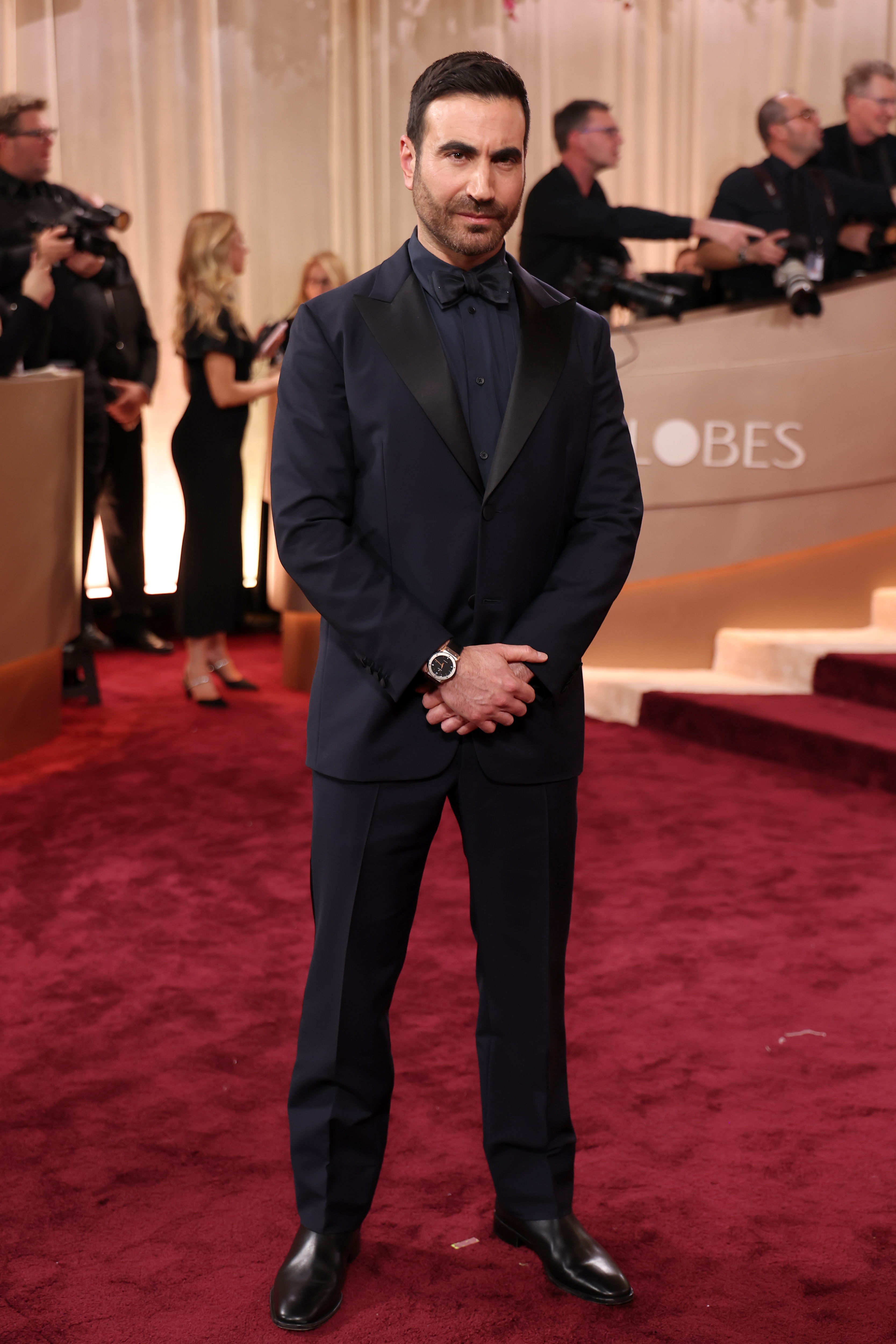 Person in a dark suit poses on the red carpet at a celebrity event, surrounded by photographers and onlookers