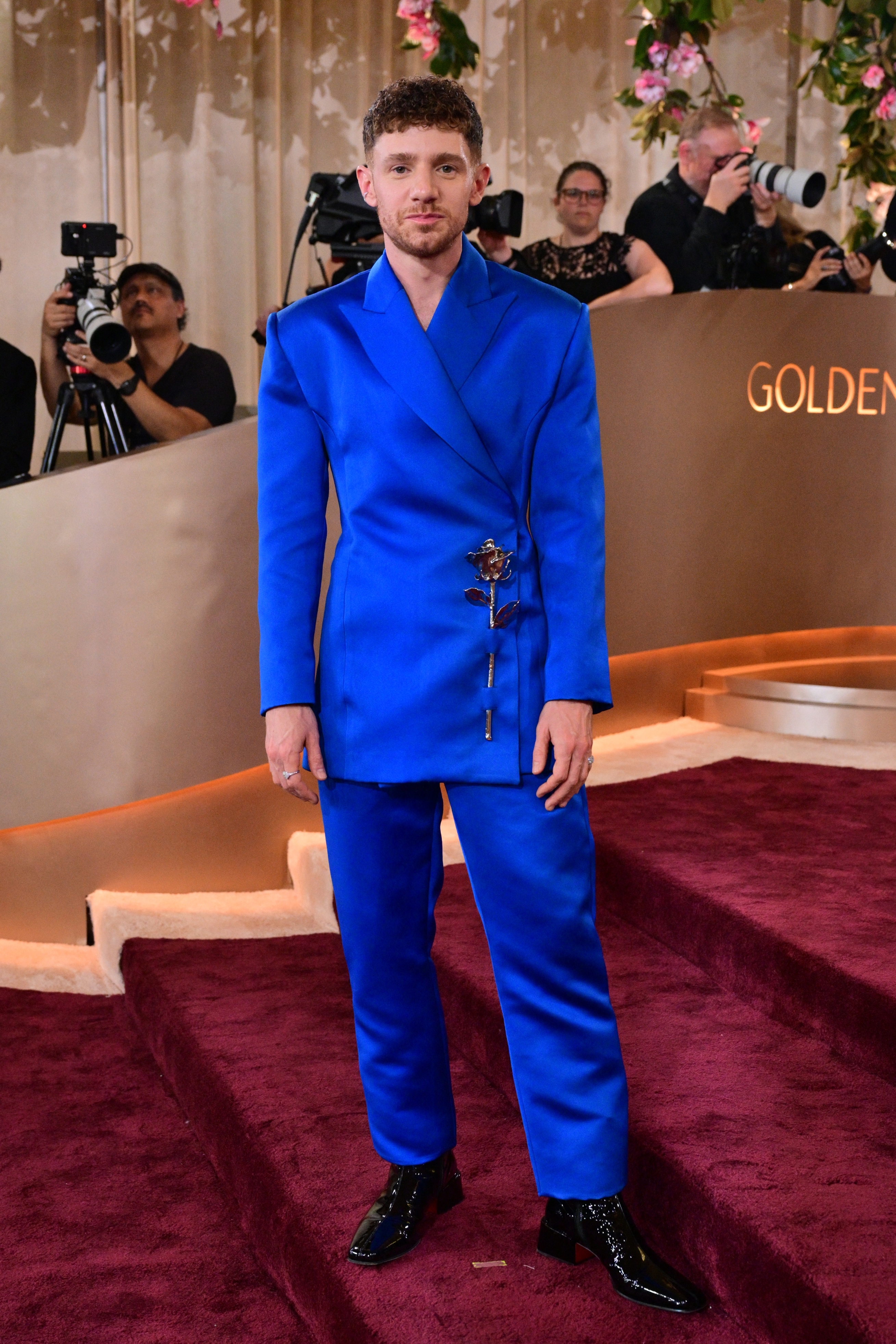 Person in a stylish blue double-breasted suit poses on a red carpet. Photographers capture the moment against a glam backdrop