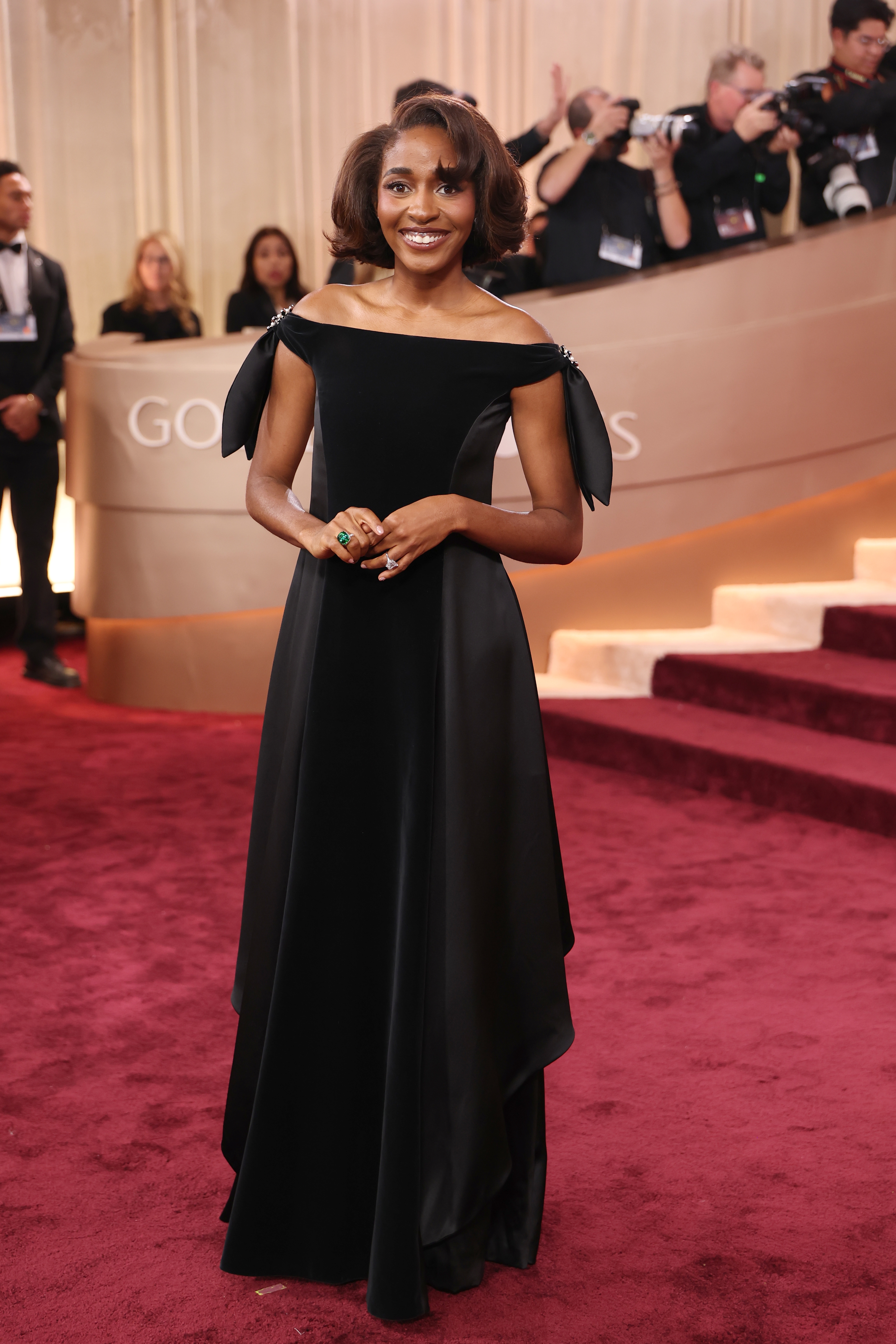 Person in an elegant, off-the-shoulder black gown poses on the red carpet at a formal event, with photographers in the background