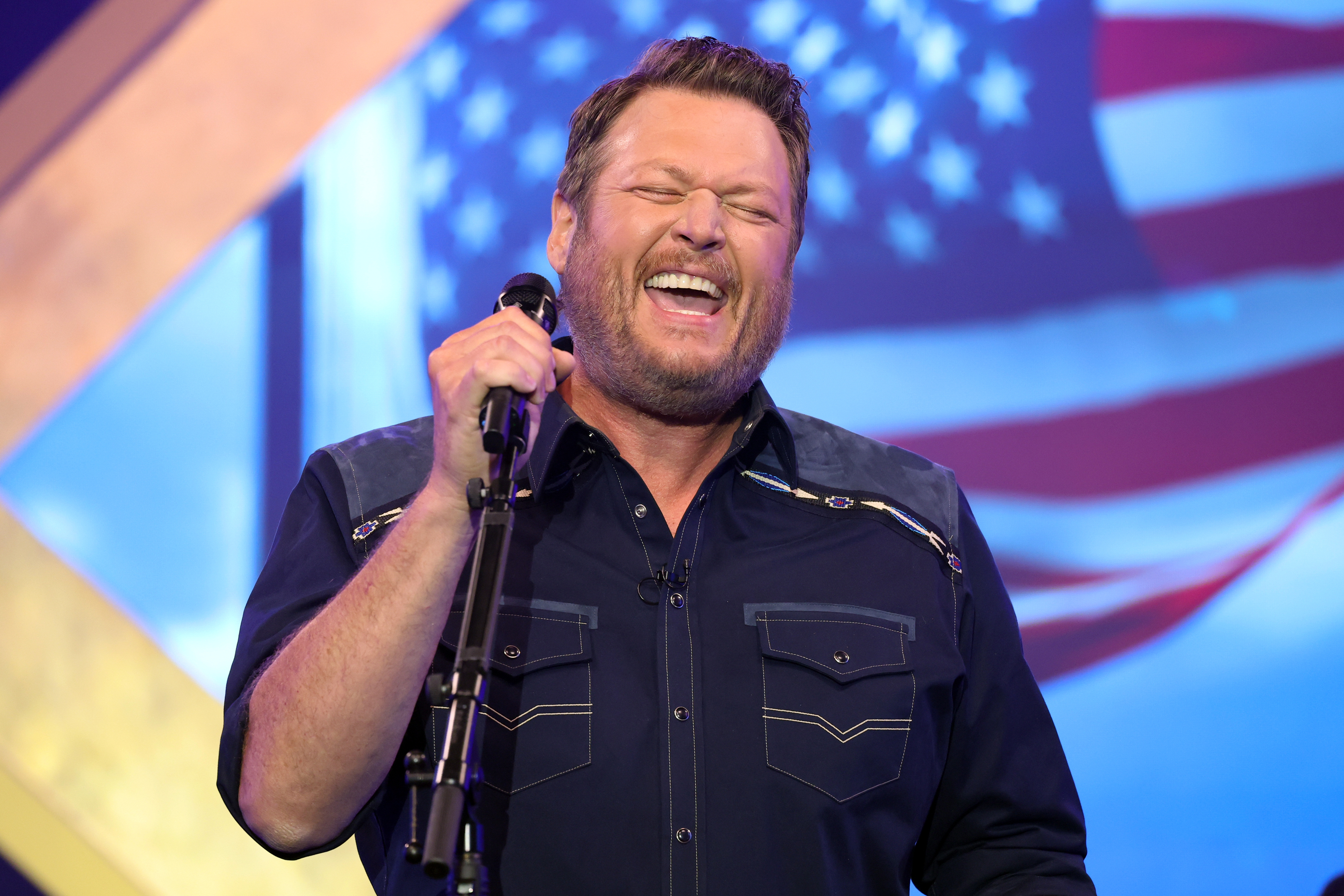 Blake Shelton singing passionately connected  stage, wearing a button-up garment  with decorative stitching, microphone successful  hand, successful  beforehand   of a blurred American emblem  backdrop