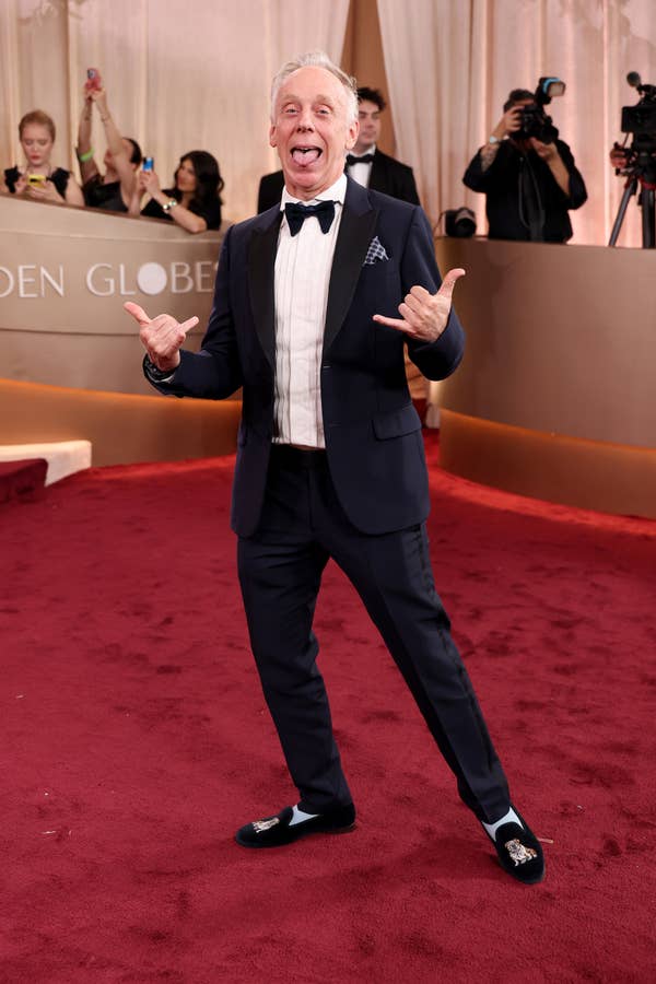 A man in a tuxedo and loafers poses playfully on the red carpet, making hand gestures with his tongue out