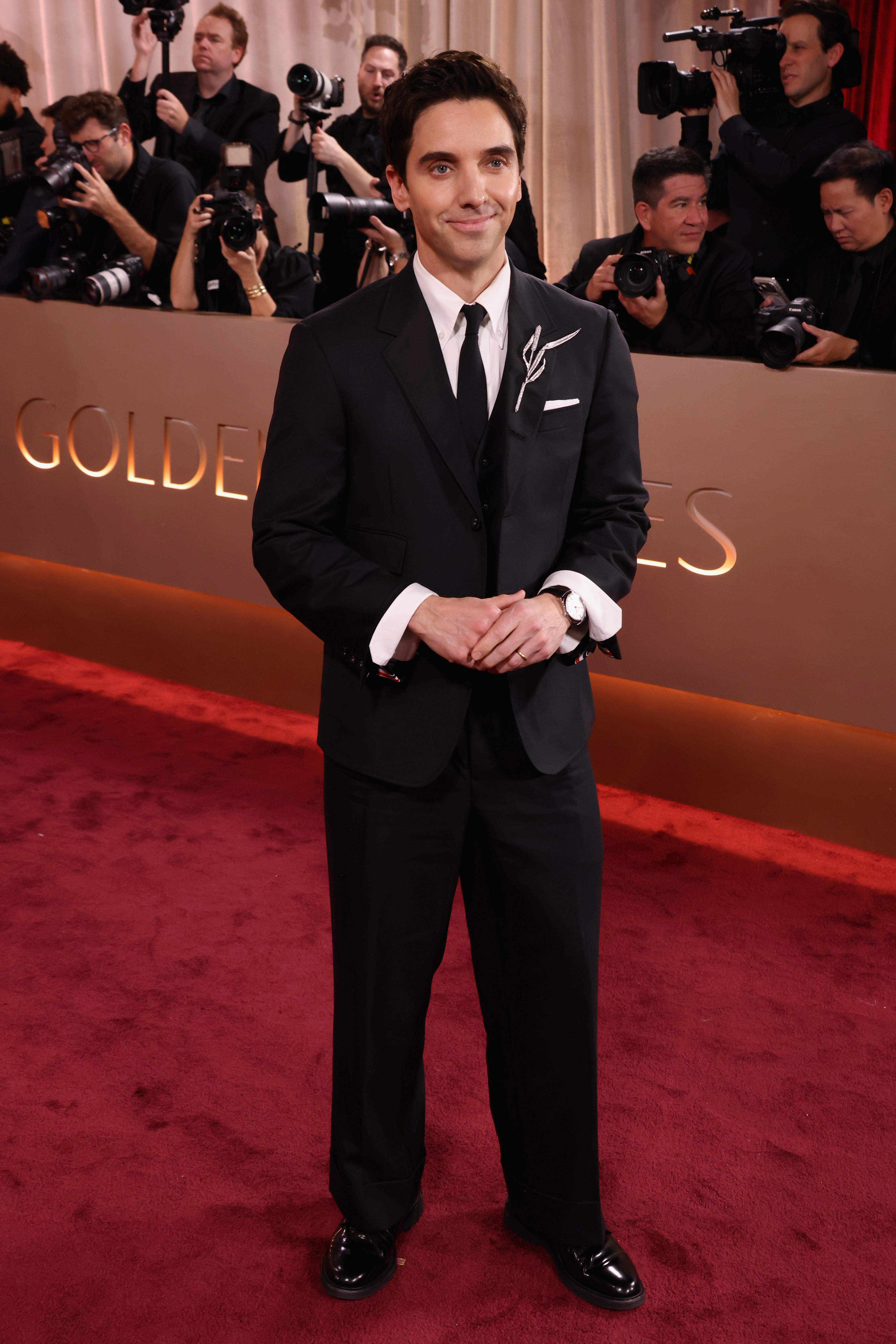 Person in a classic black tuxedo poses on the red carpet with photographers in the background