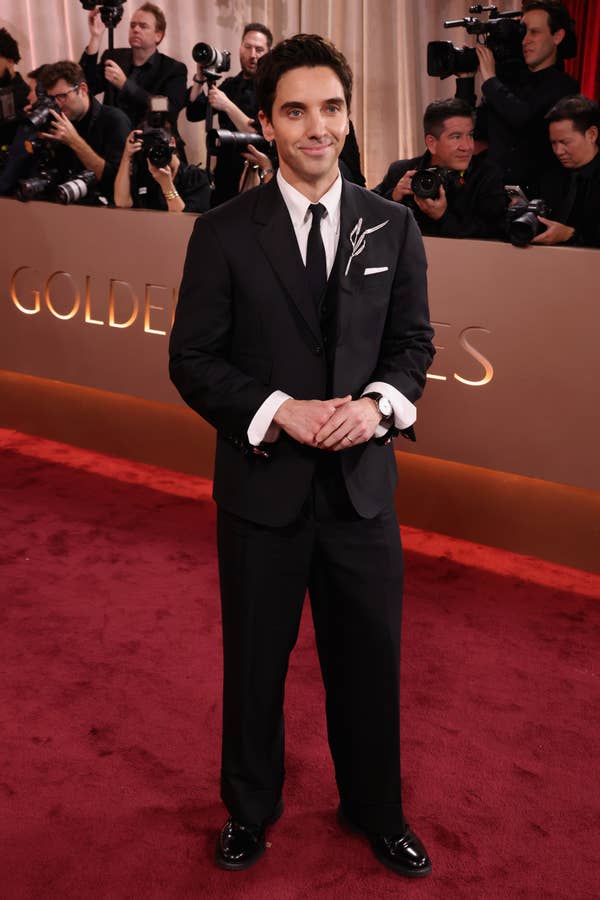 Person in a classic black tuxedo poses on the red carpet with photographers in the background