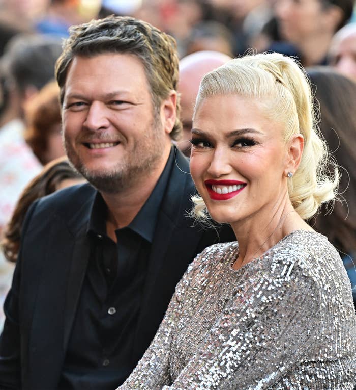 Gwen Stefani and Blake Shelton smiling astatine  an event. One wears a acheronian  suit, the different   a sparkling outfit. Crowd seen successful  the background