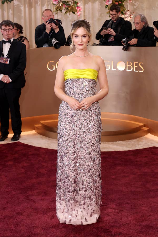 A person poses on the red carpet in a strapless gown with a textured skirt and bold top, while photographers capture the moment in the background