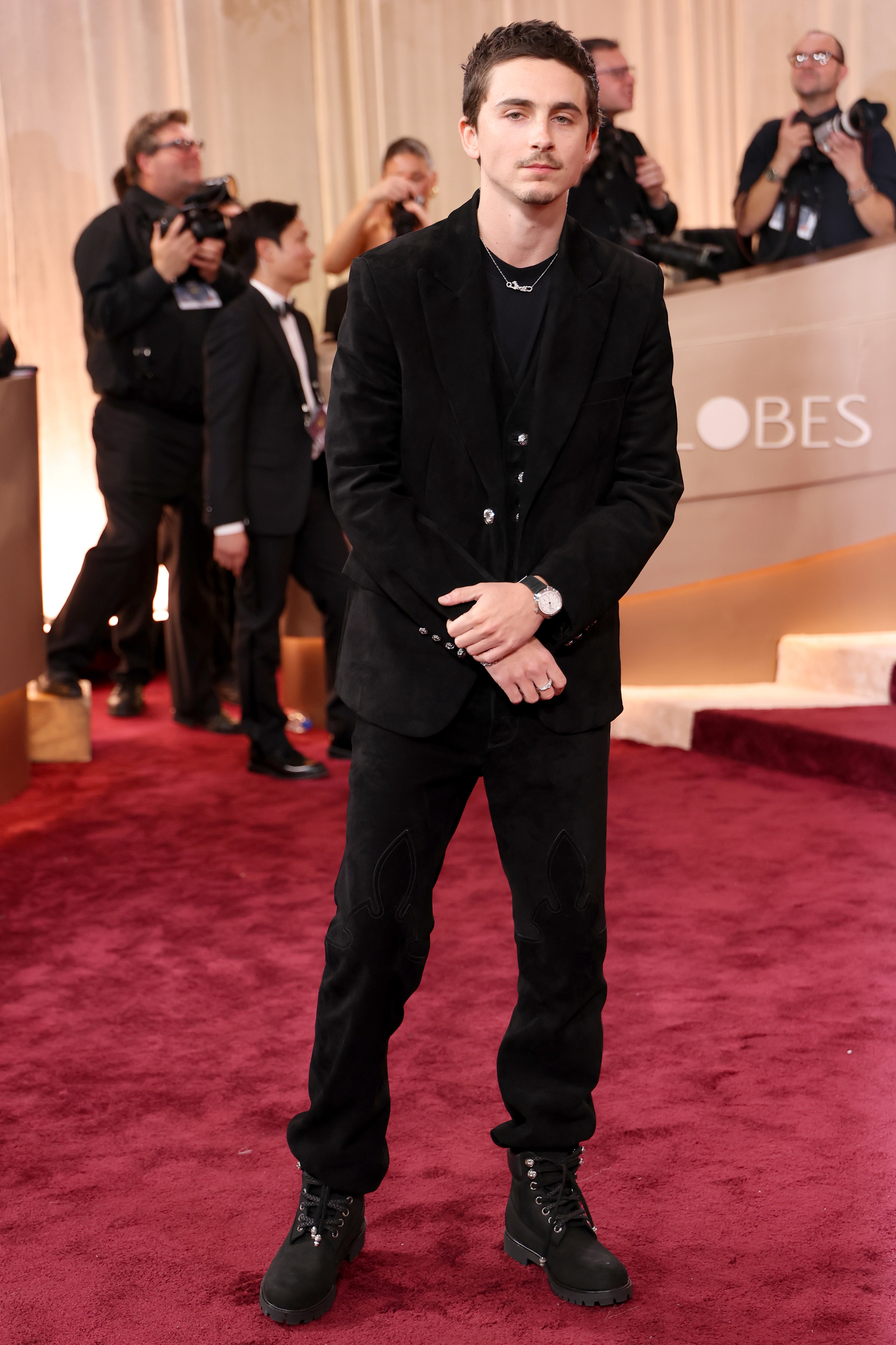 Person in tailored black suit and boots on a red carpet with photographers in the background