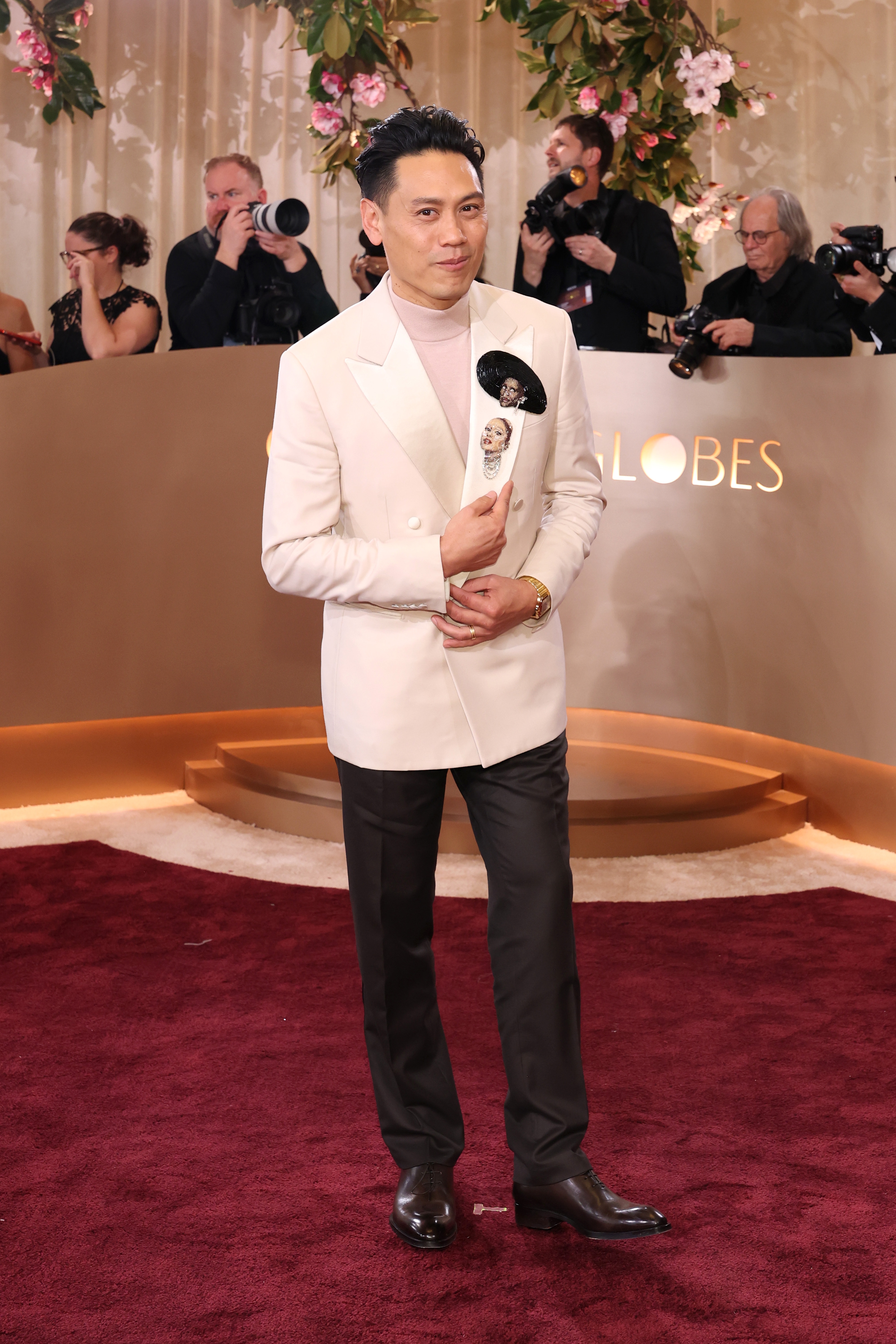 A man on the red carpet at a celebrity event, wearing a white double-breasted blazer and dark pants, holds a fan with a playful face design