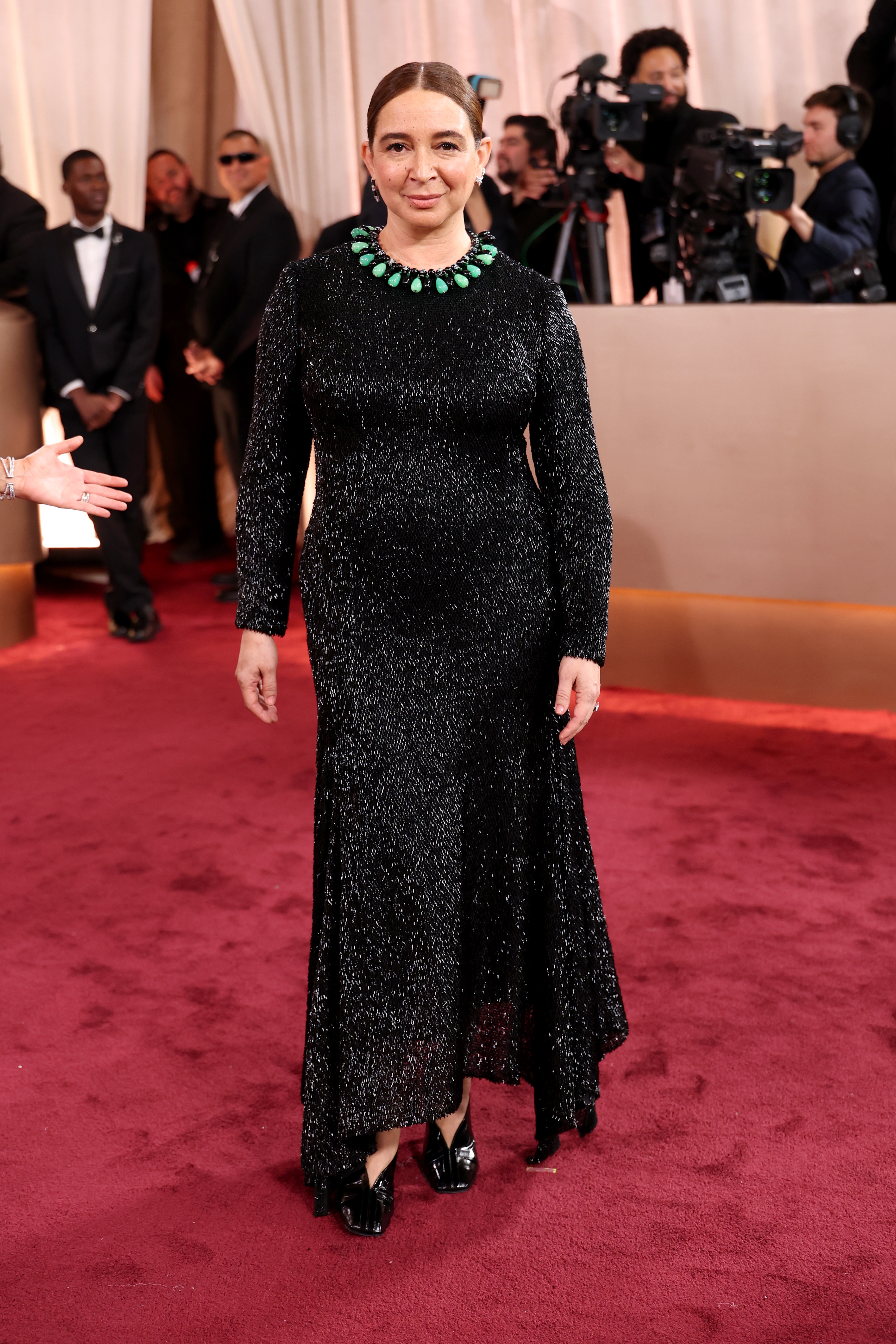 Person in a sparkling black gown with a green necklace on a red carpet. Camera crew seen in the background