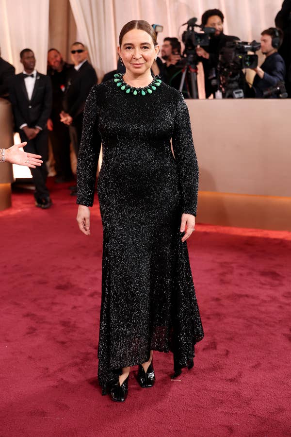 Person in a sparkling black gown with a green necklace on a red carpet. Camera crew seen in the background