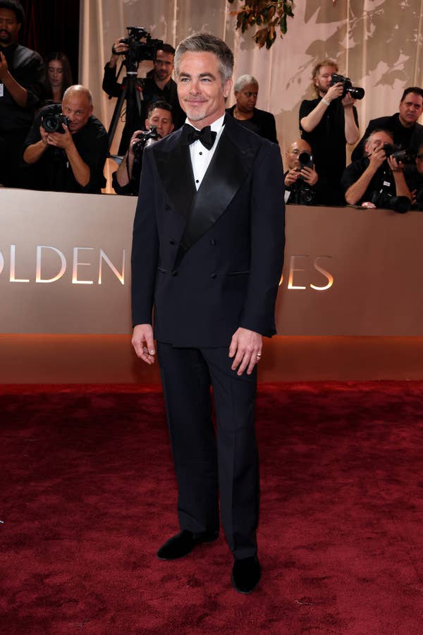 Celebrity in formal suit with black lapels poses on a red carpet, surrounded by photographers at a high-profile event