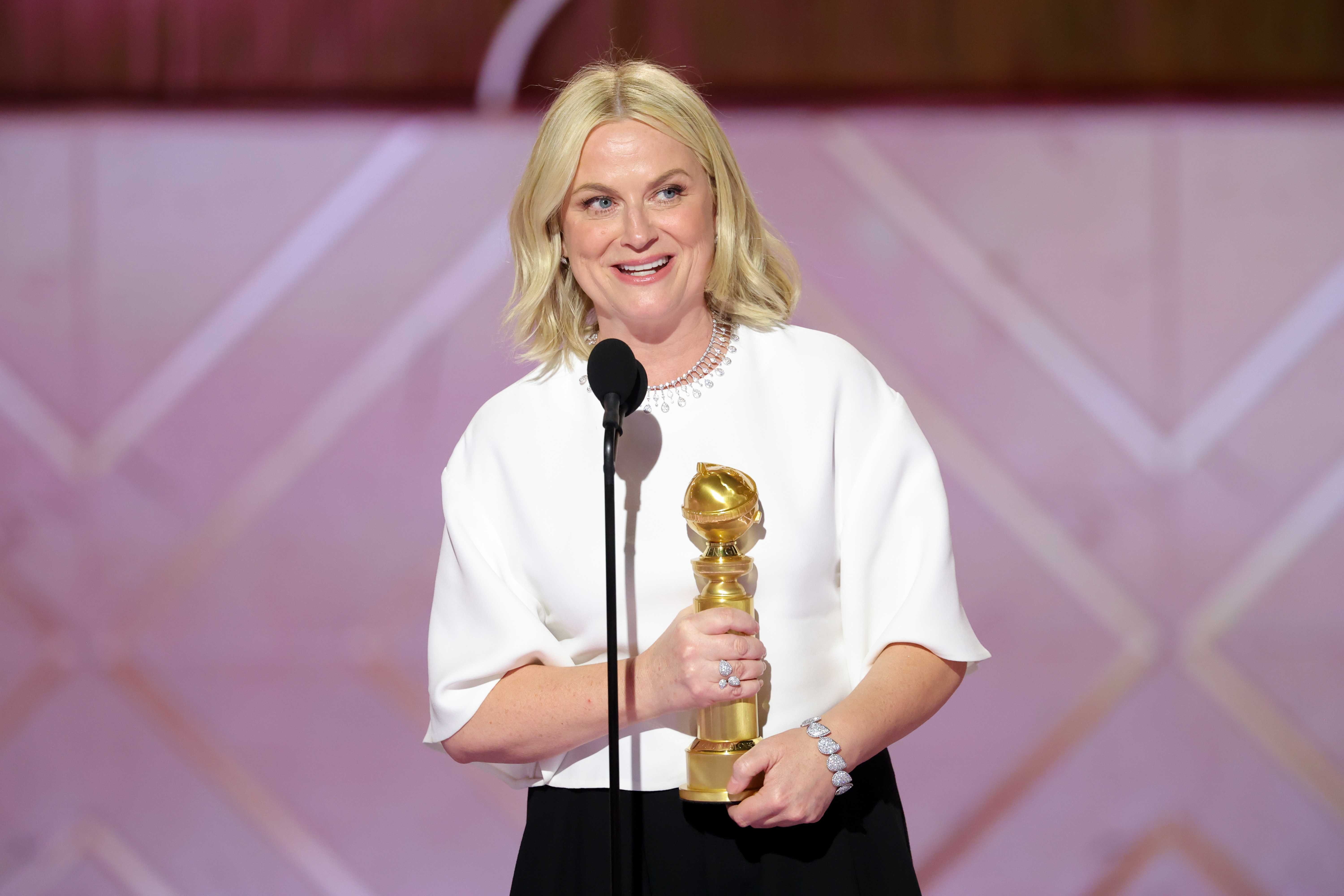 Person som holder et Golden Globe-trofé, iført en elegant hvit topp med rund hals, snakker ved et podium