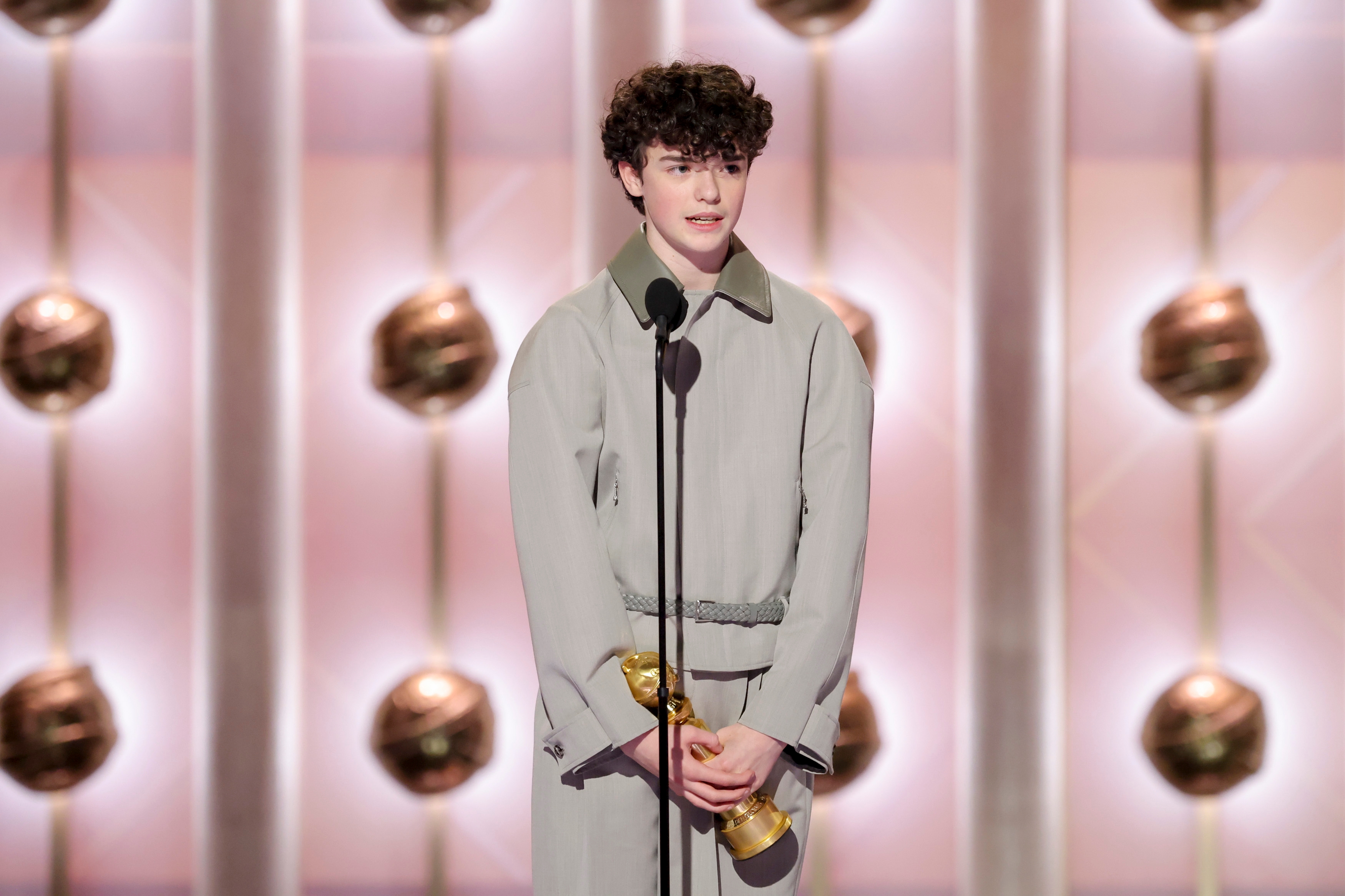 Person i en stilig lang frakk som holder et trofé på scenen, snakker i en mikrofon mot et bakteppe med hengende dekorasjoner