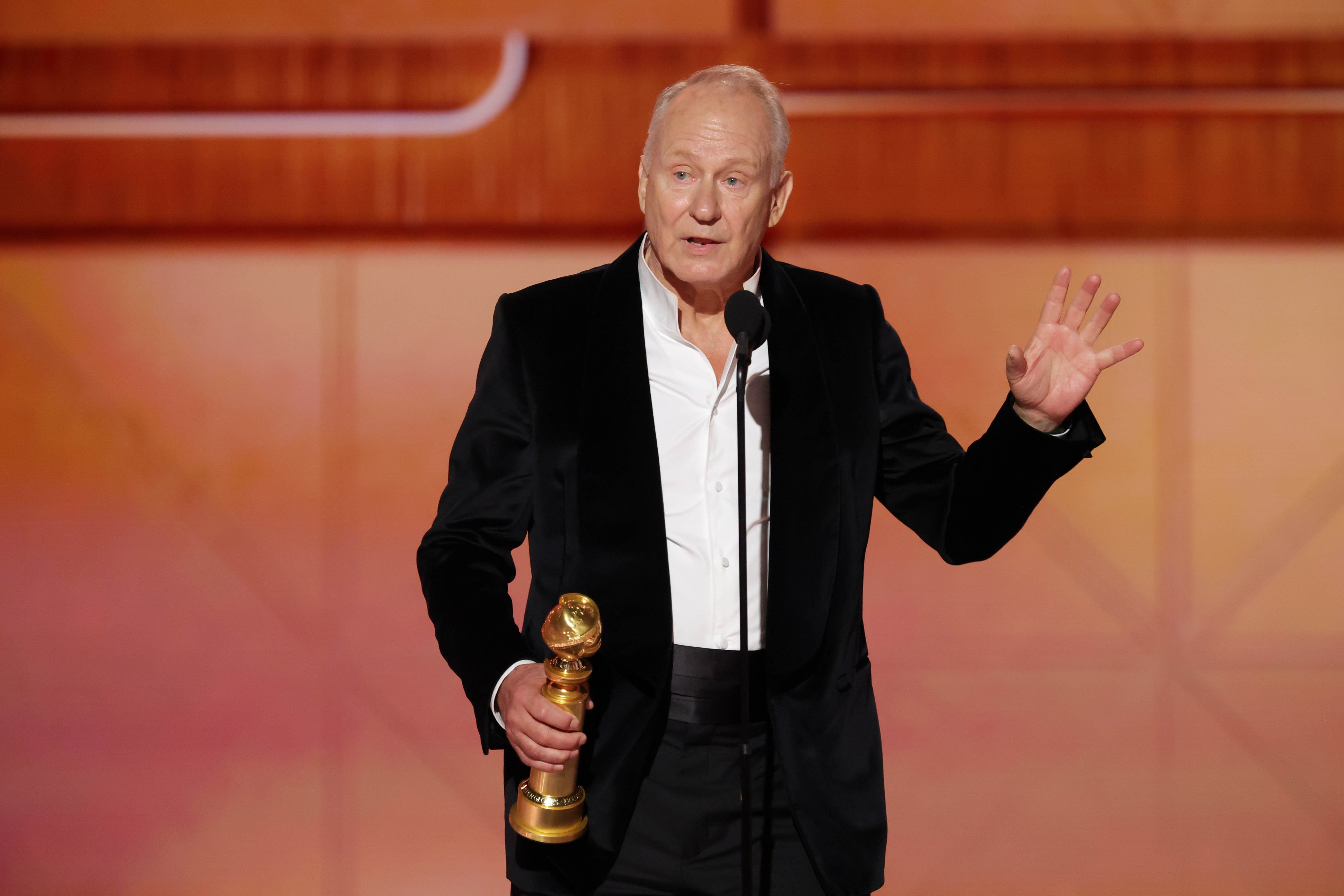 En person i svart dress og hvit skjorte taler på scenen med et gullpokal