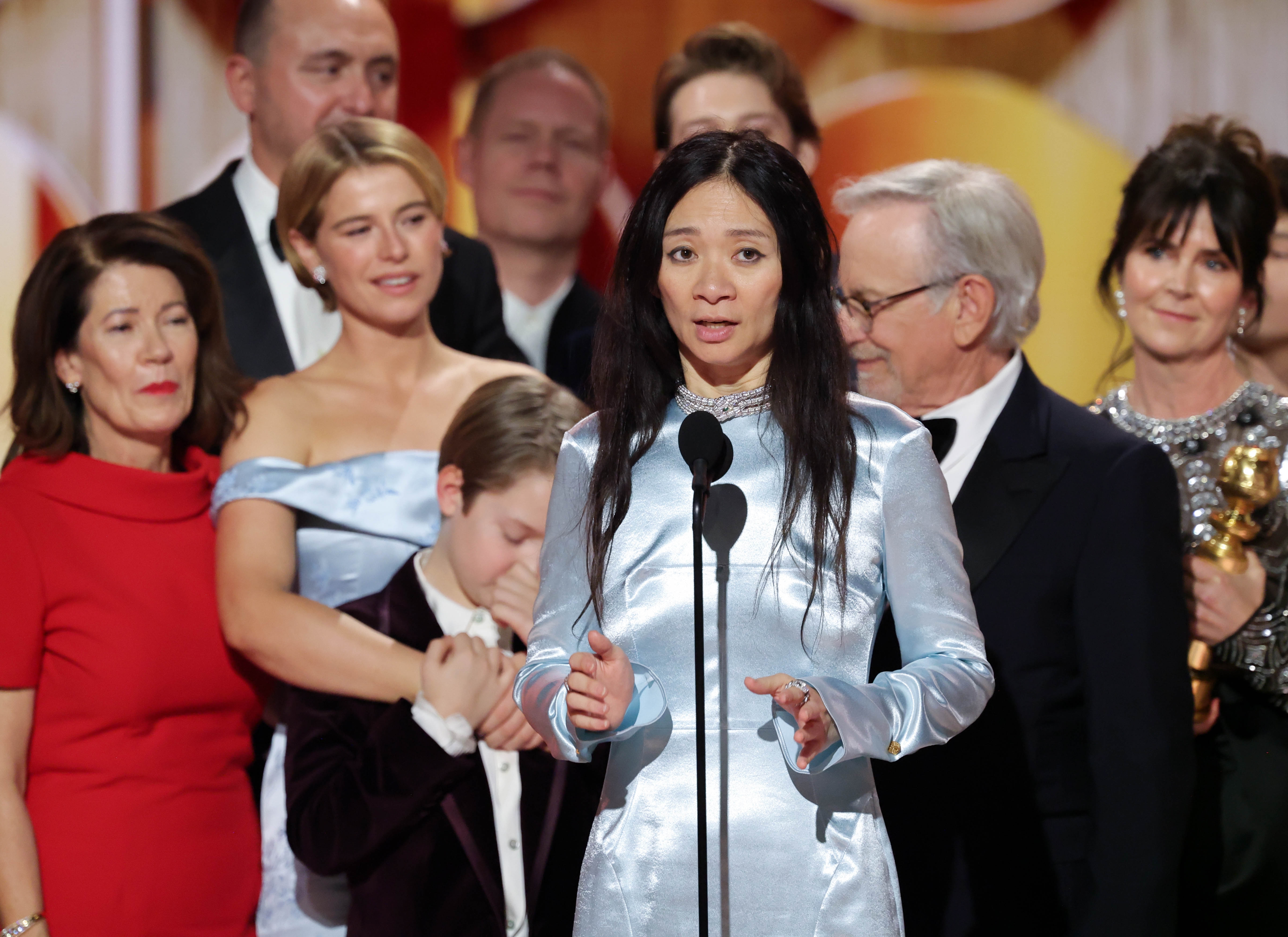 En gruppe mennesker på scenen under en prisutdeling, en kvinne i en satengkjole snakker inn i en mikrofon. Andre står støttende bak henne