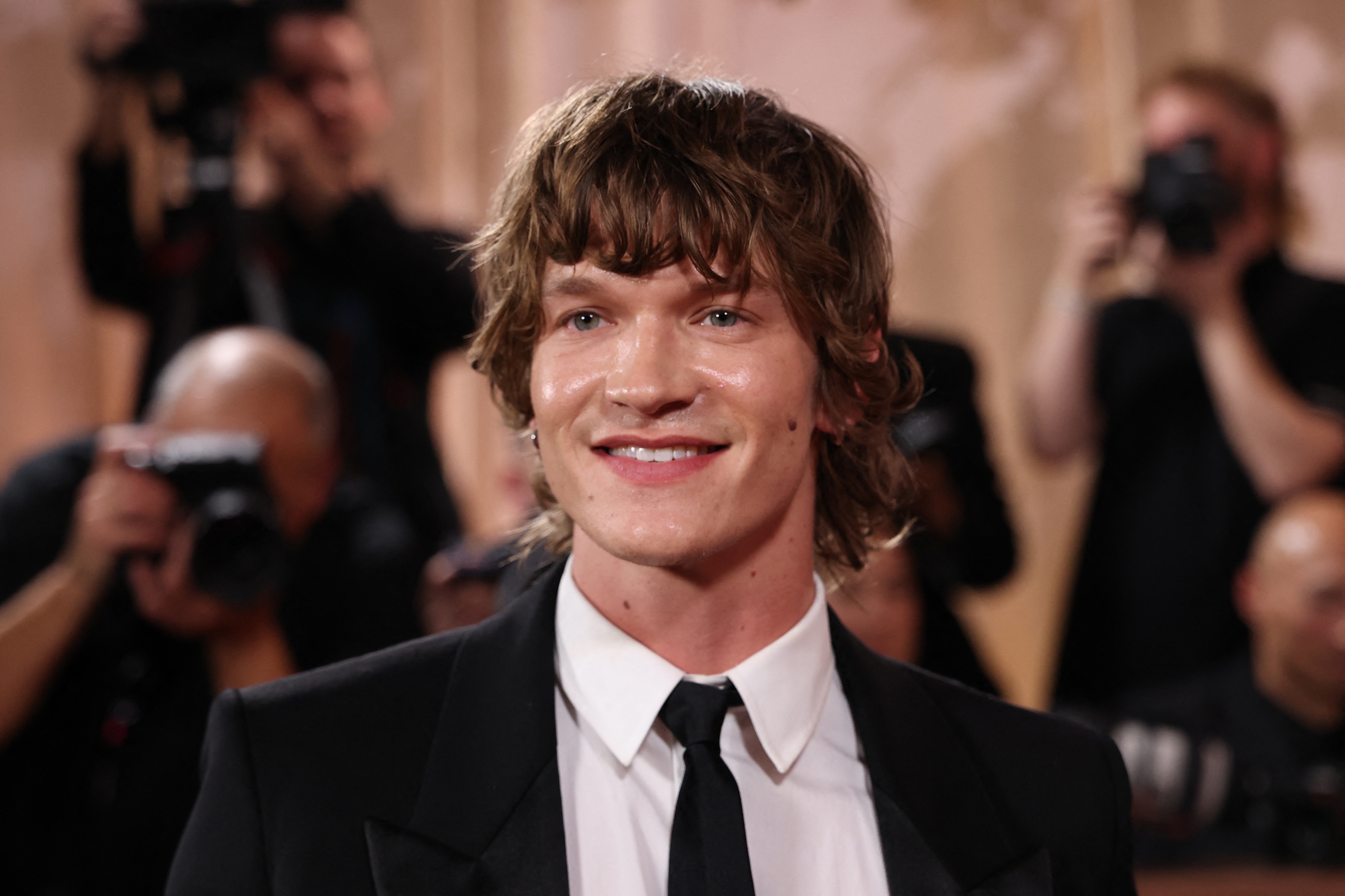 Smilende person i dress og slips på en rød løper, omgitt av fotografer