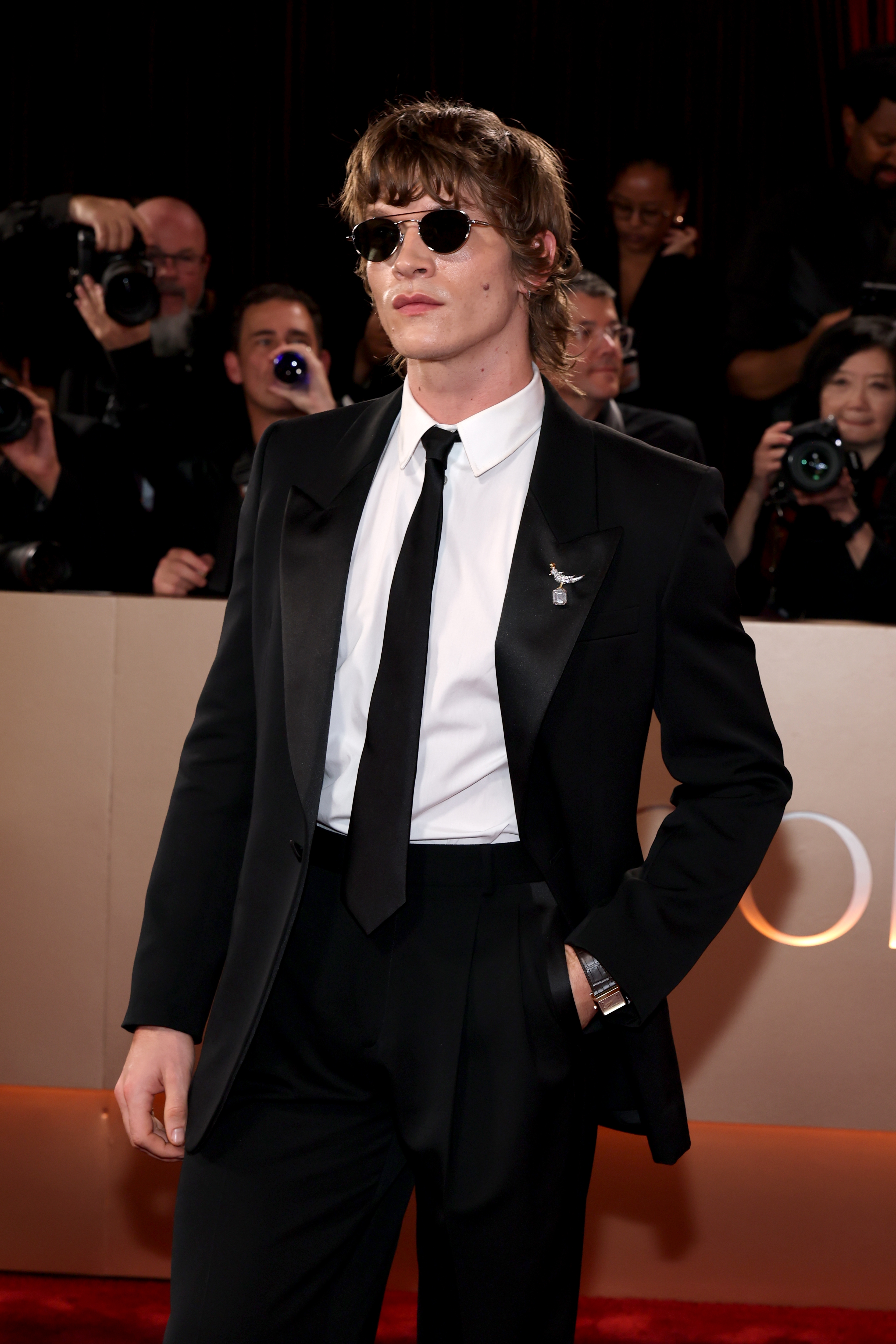 Person i klassisk svart smoking og slips, med solbriller, poserer selvsikkert på en rød løper med fotografer i bakgrunnen