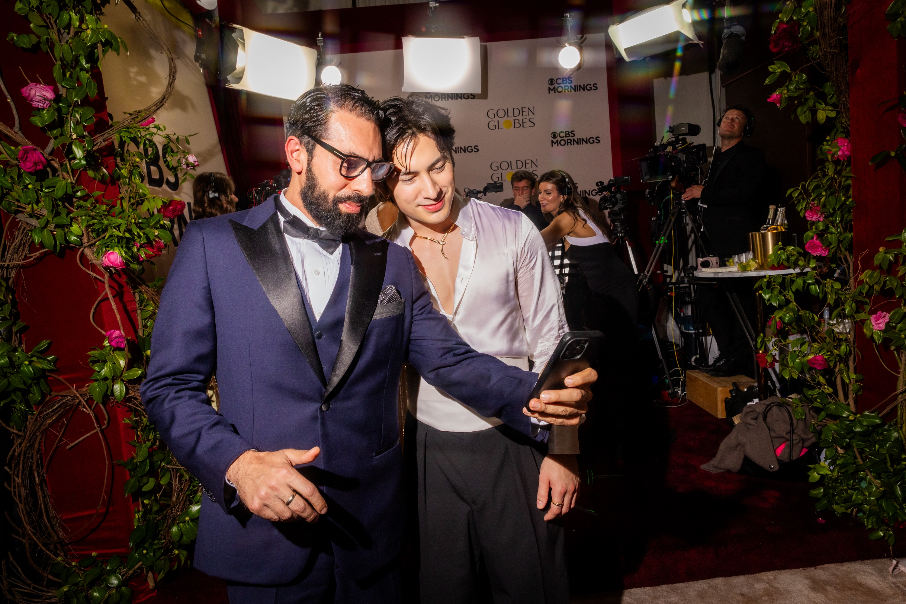 To personer tar en selfie på en rød løper. Man har på seg en blå smoking med sløyfe; den andre har på seg en silkehvit skjorte med lav utringning