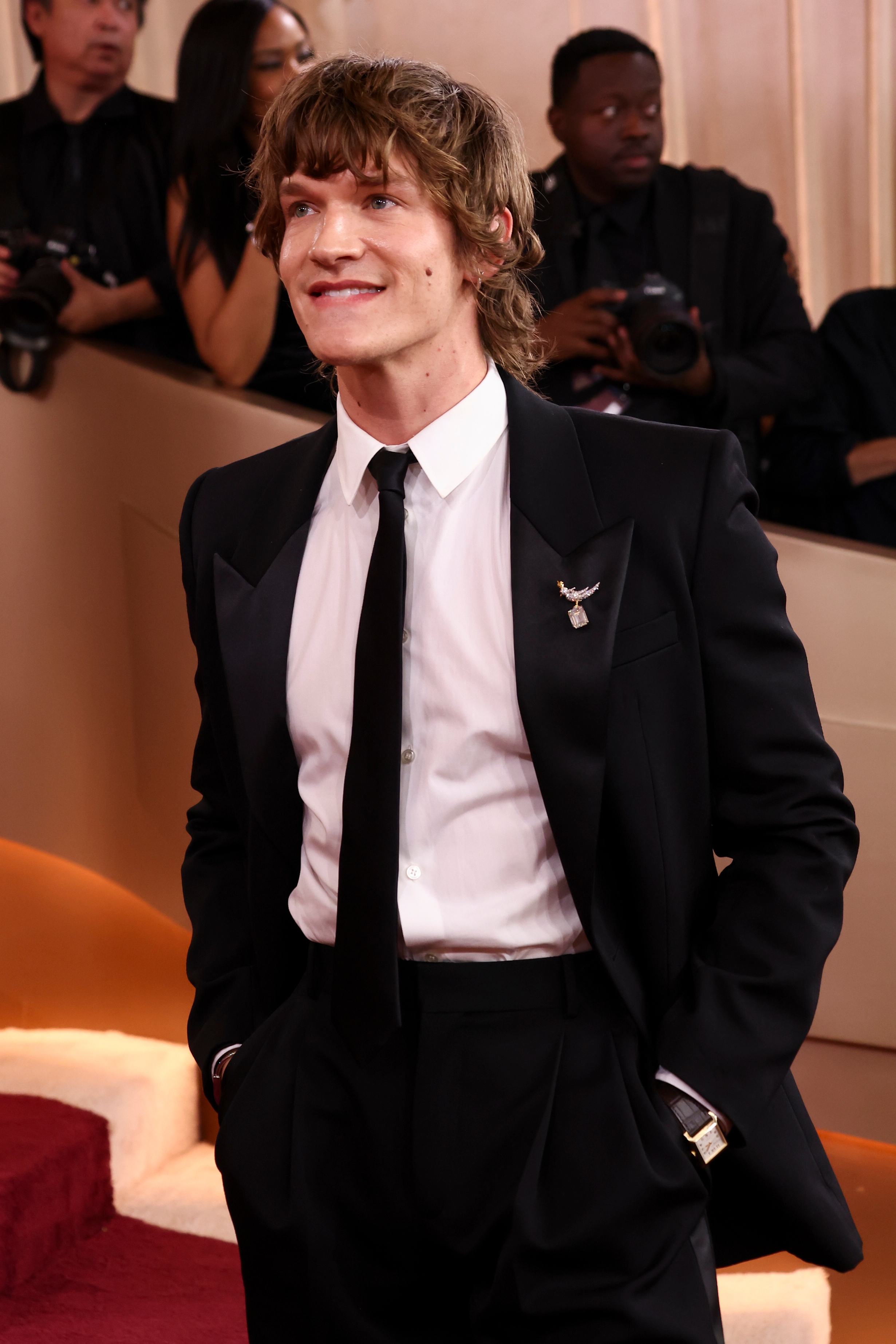 Person i skreddersydd svart dress med slips, smilende på den røde løperen. Andre deltakere er i bakgrunnen