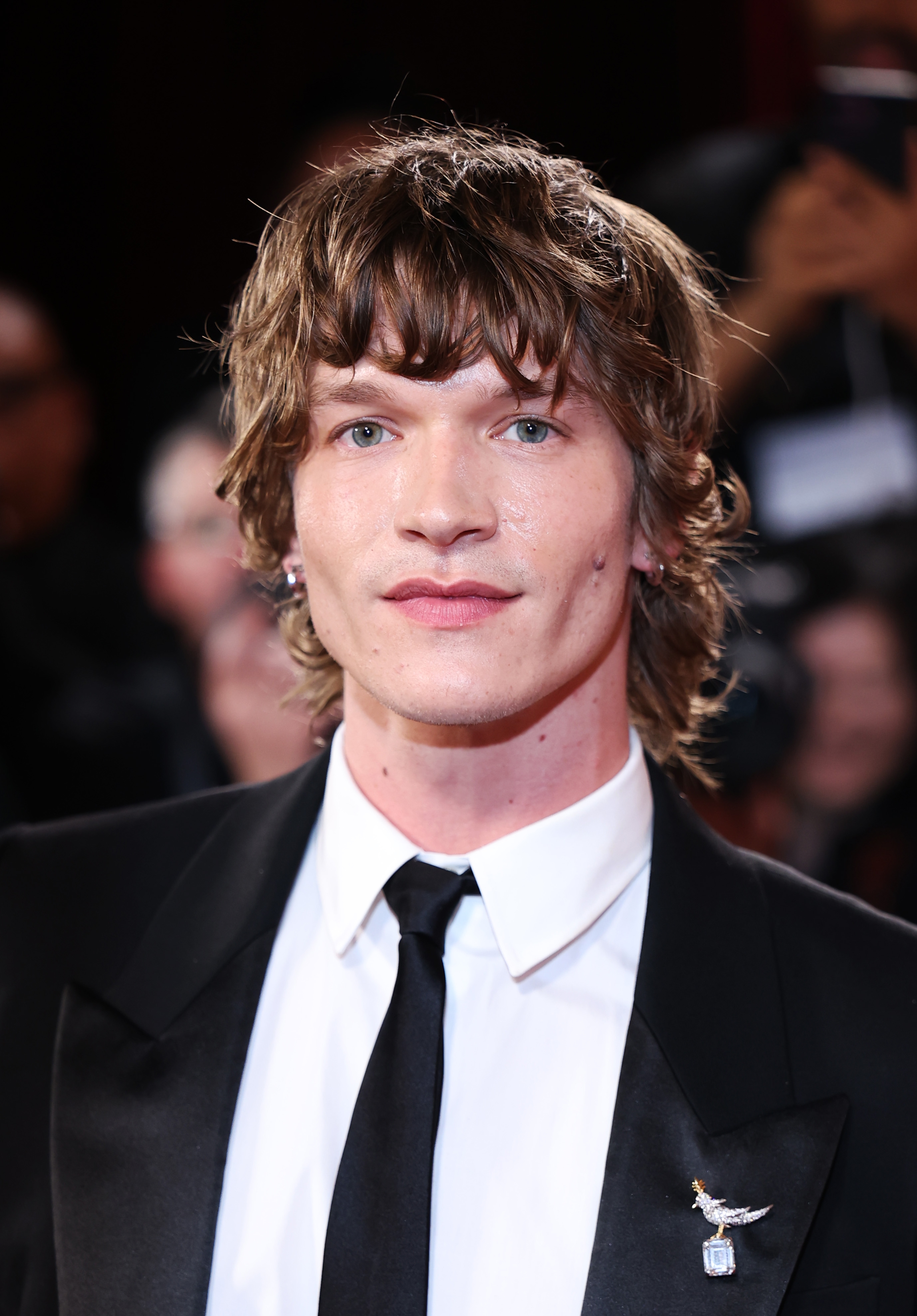 Person i en formell dress på en rød løper, iført en jakkeslagsnål, med bølget hår og et nøytralt uttrykk