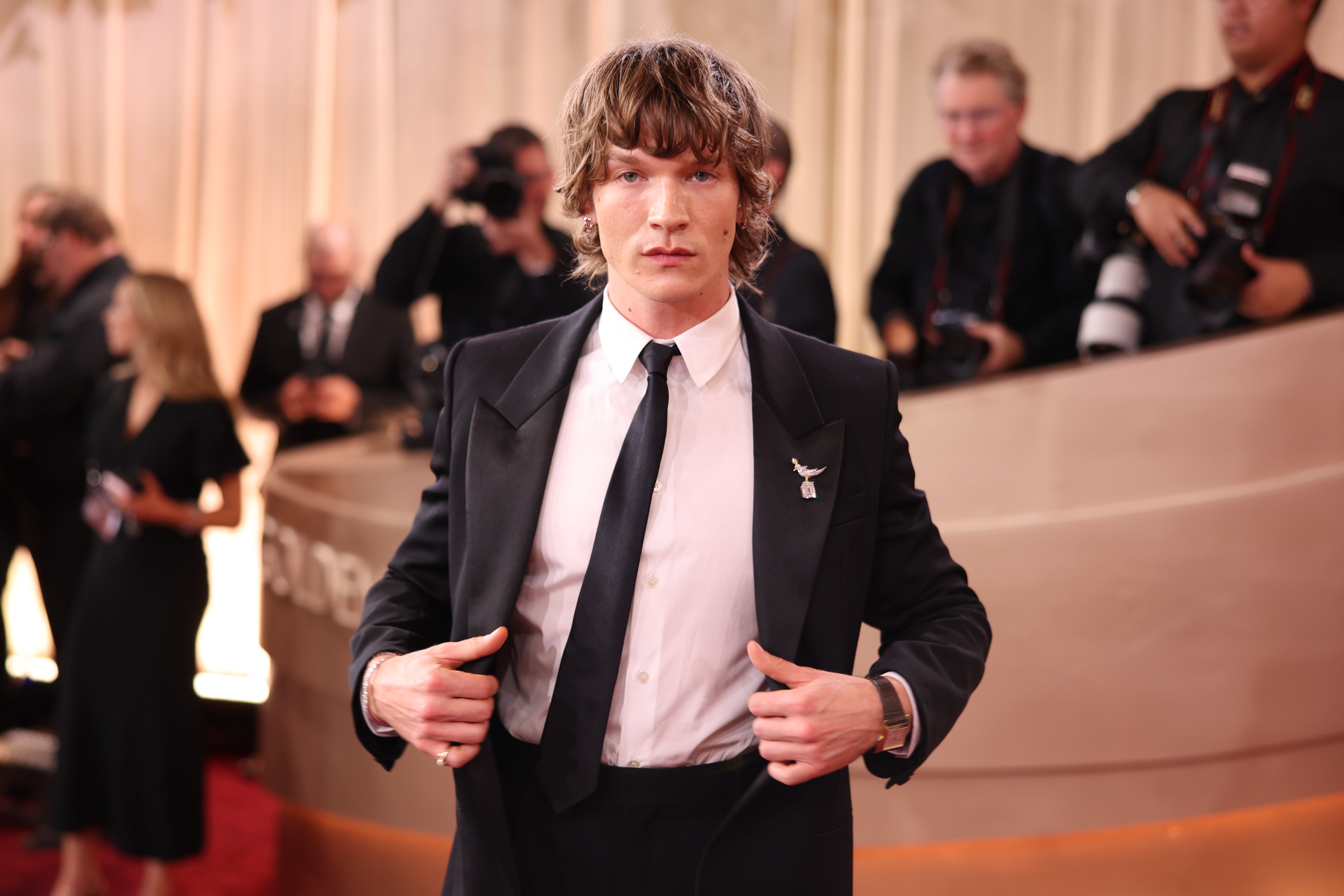 Person i en skreddersydd svart dress med slips på en rød løper, fotografer i bakgrunnen