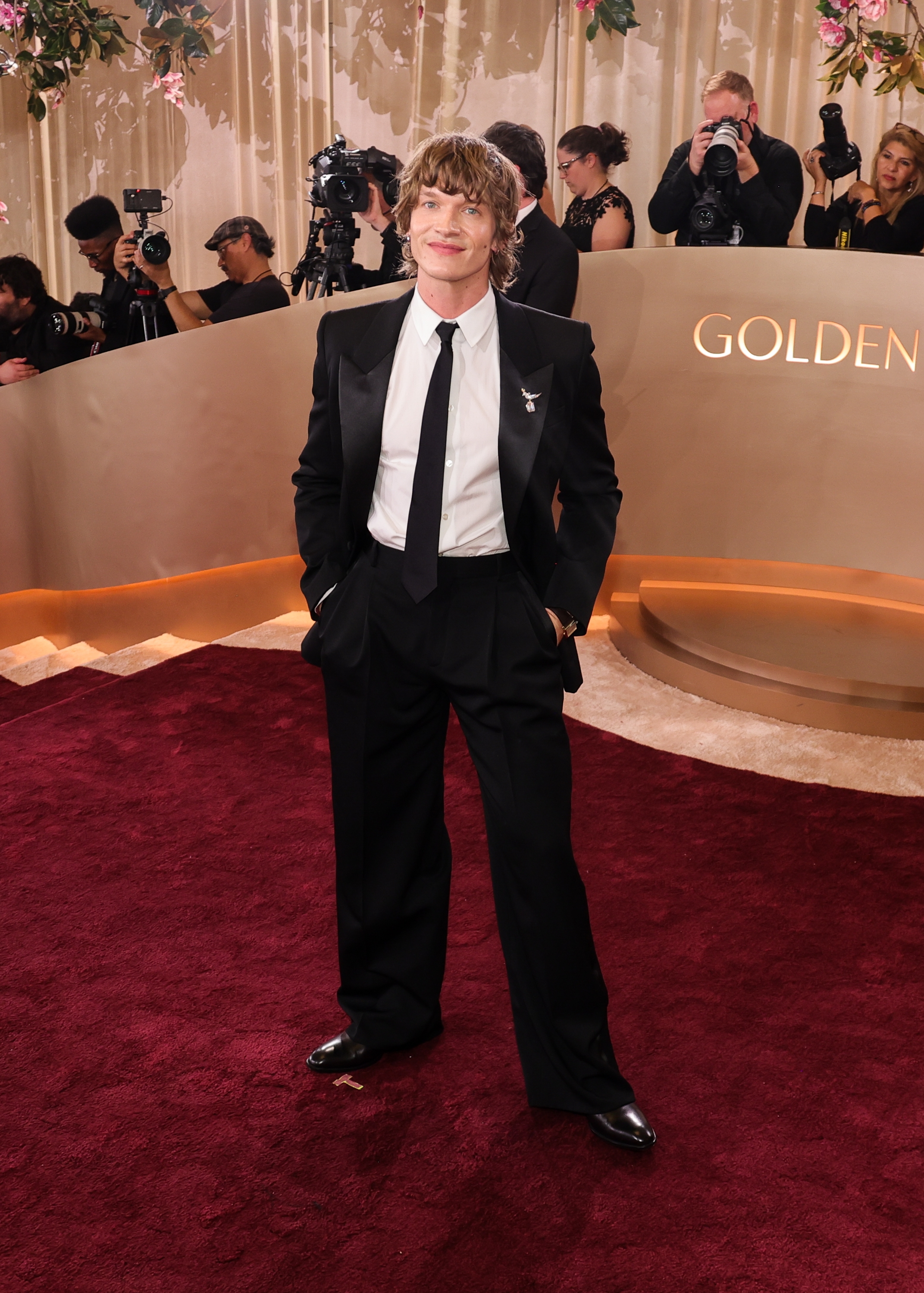 Person i en formell svart dress med slips står på en rød løper og poserer for bilder på et arrangement med fotografer i bakgrunnen