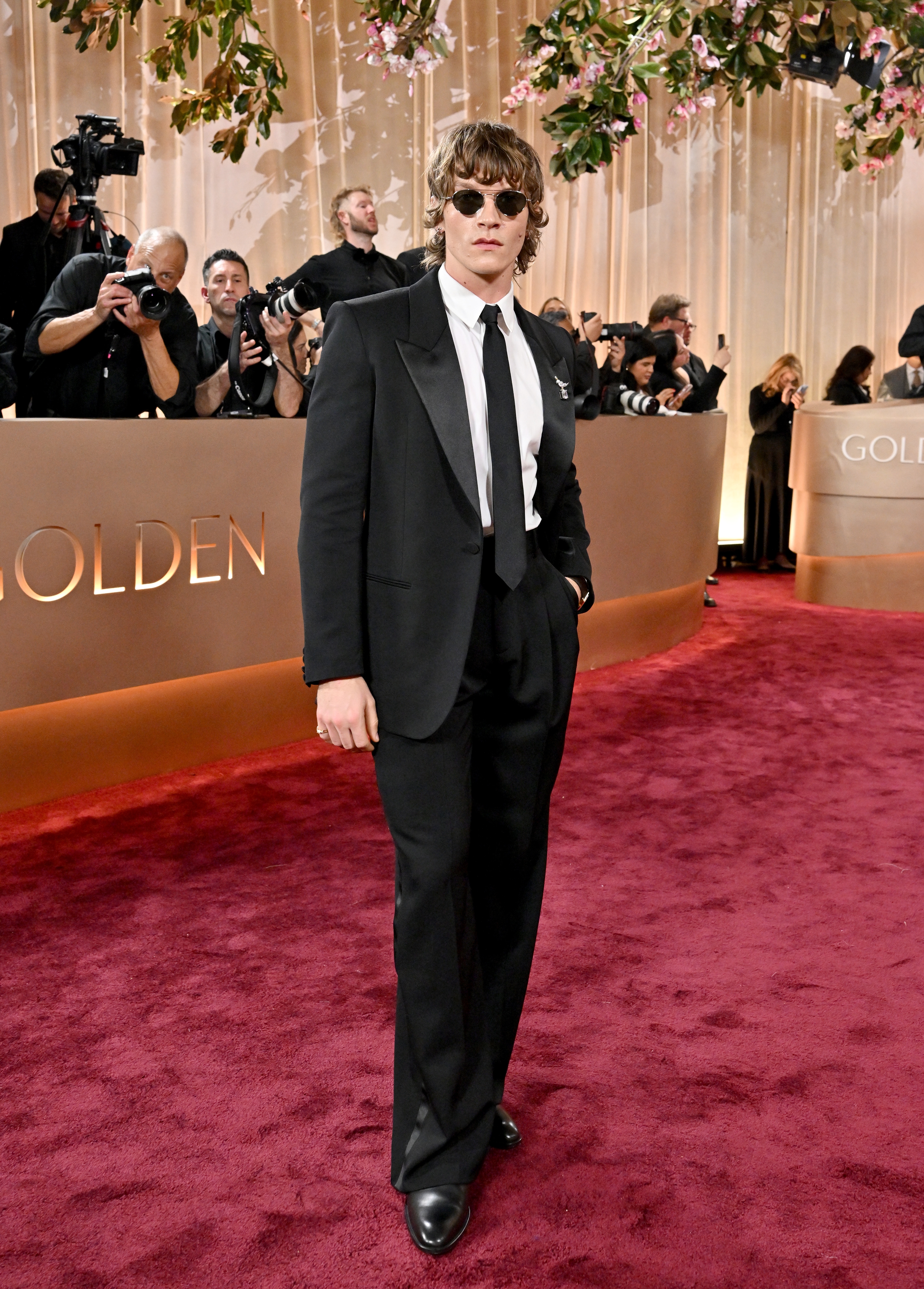 Person i en formell svart dress med solbriller på en rød løper, fotografer i bakgrunnen