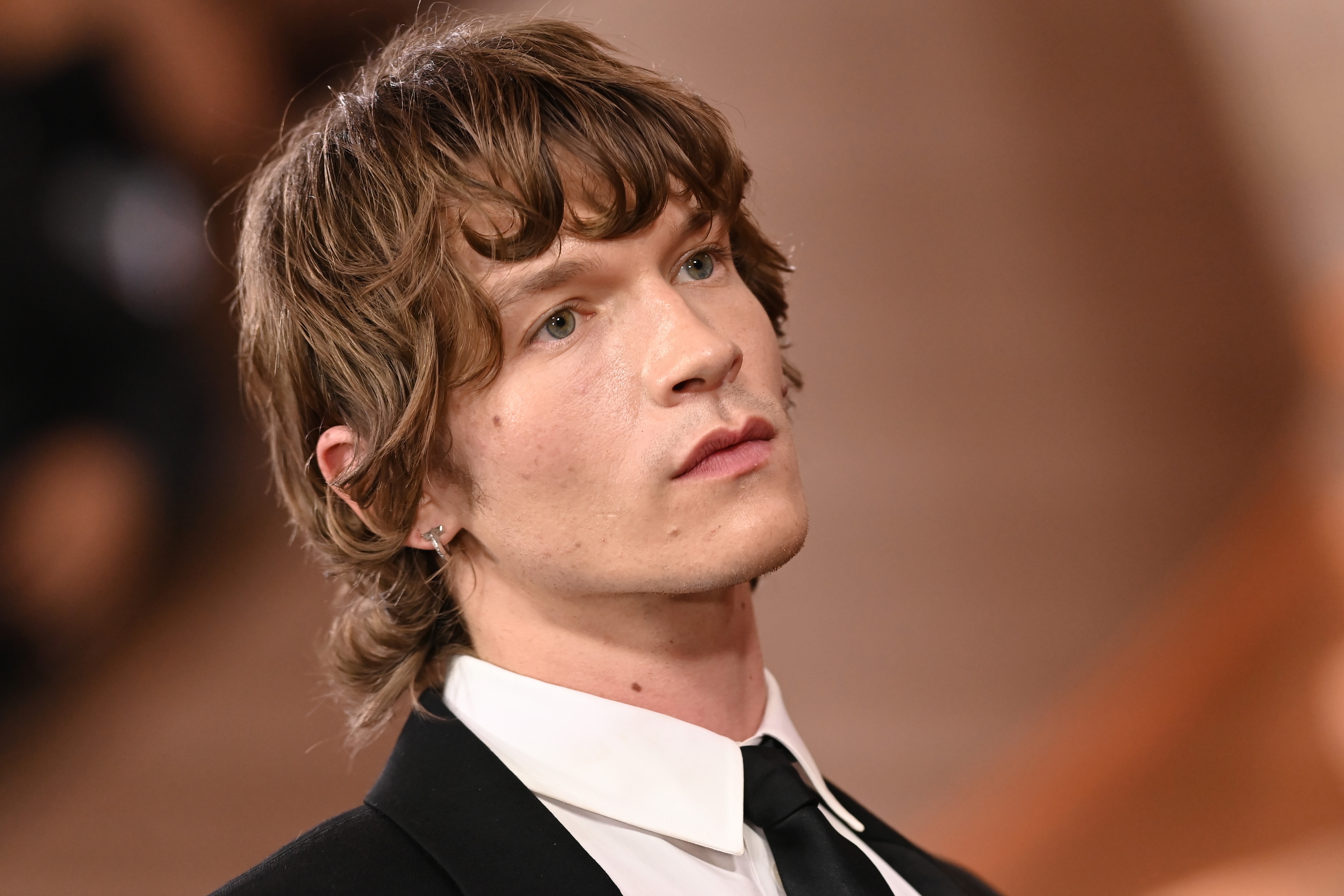 Person i formell antrekk med bølget hår på en rød løper, iført svart dress og hvit skjorte, ser oppover