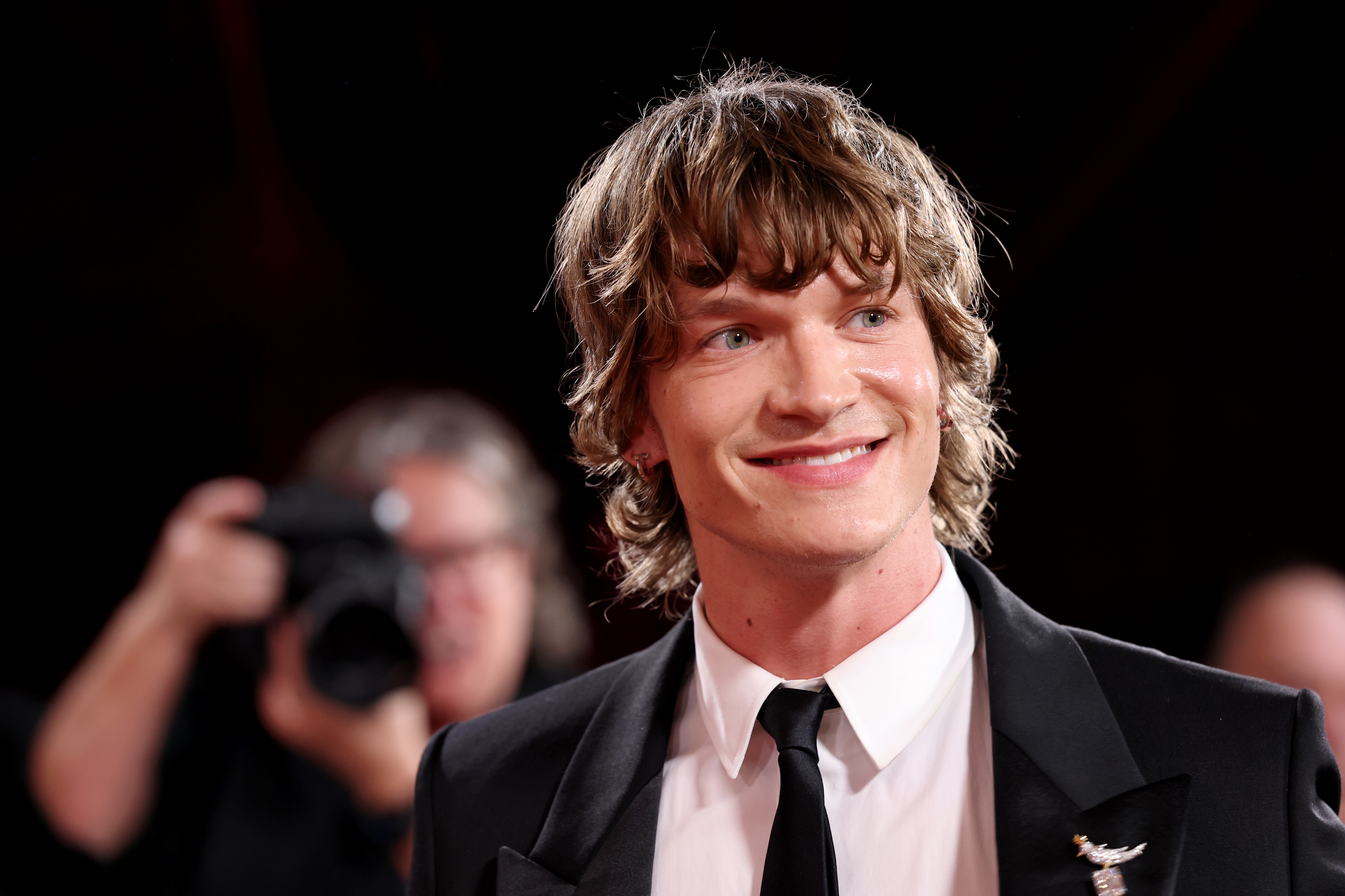 Person i klassisk svart dress og slips smiler på en rød løper, med fotografer synlige i bakgrunnen