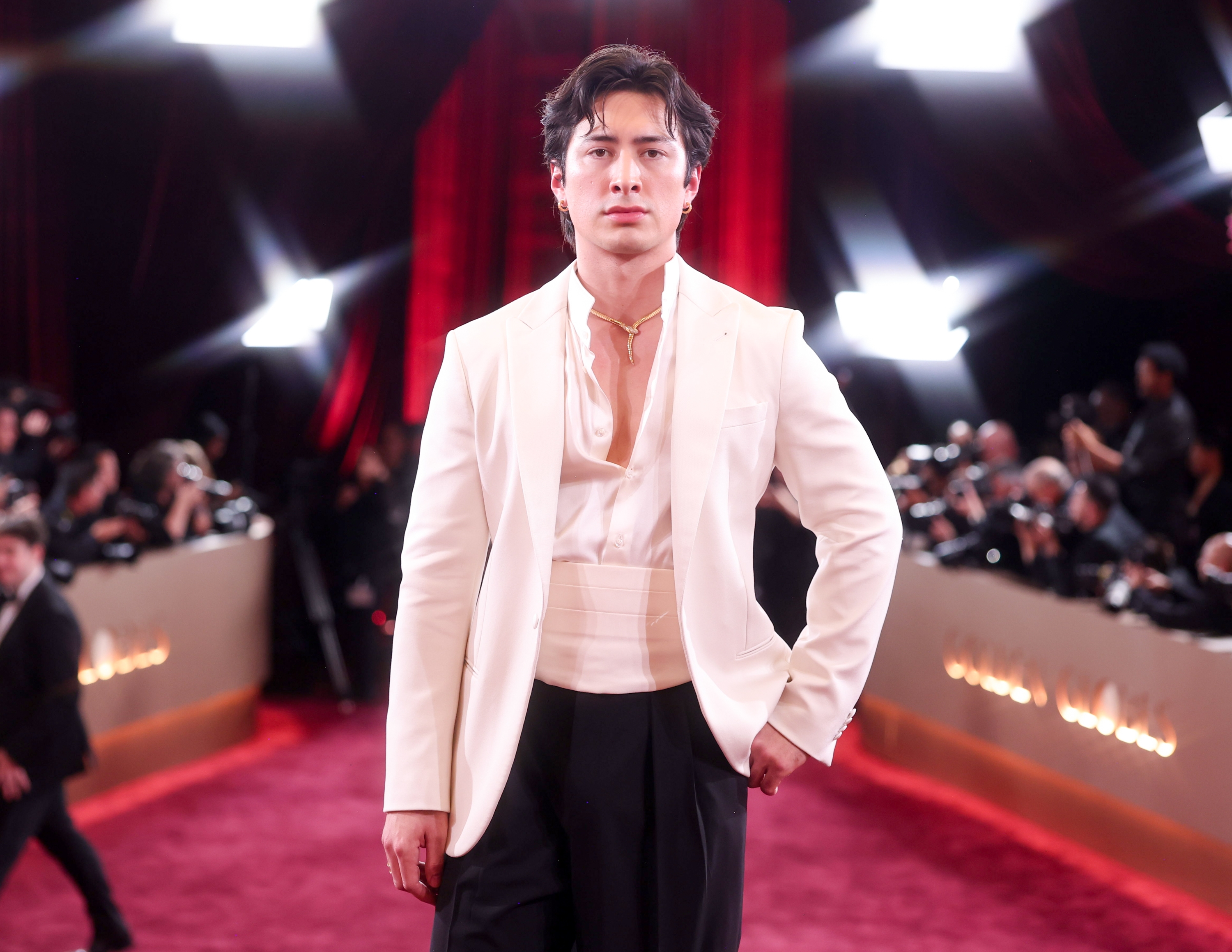 Person på rød løper iført en hvit blazer, skjorte med åpen krage og en cummerbund med svarte bukser, omgitt av fotografer