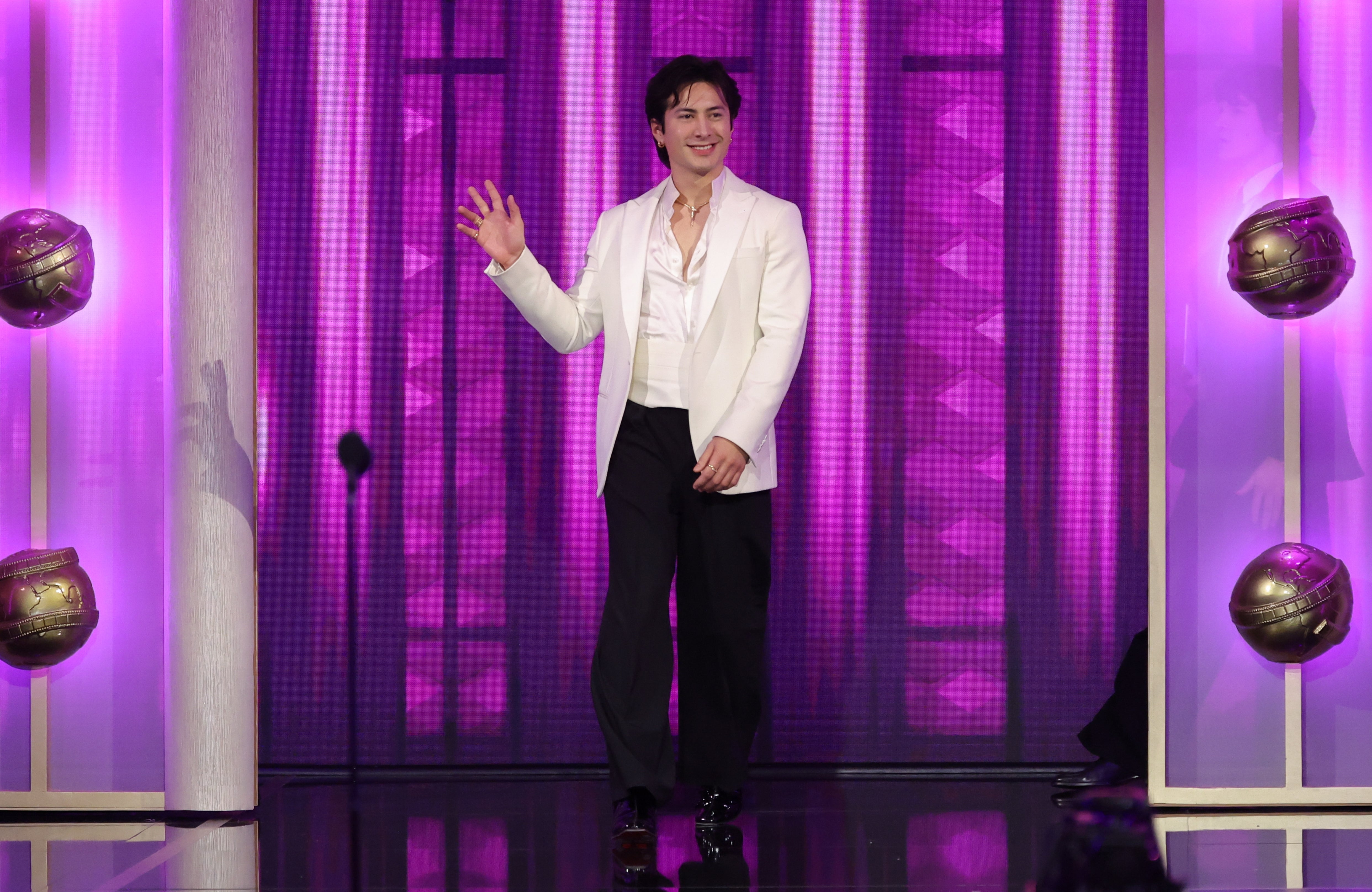 Person i hvit jakke og svarte bukser vinker på scenen med lilla opplyst bakgrunn