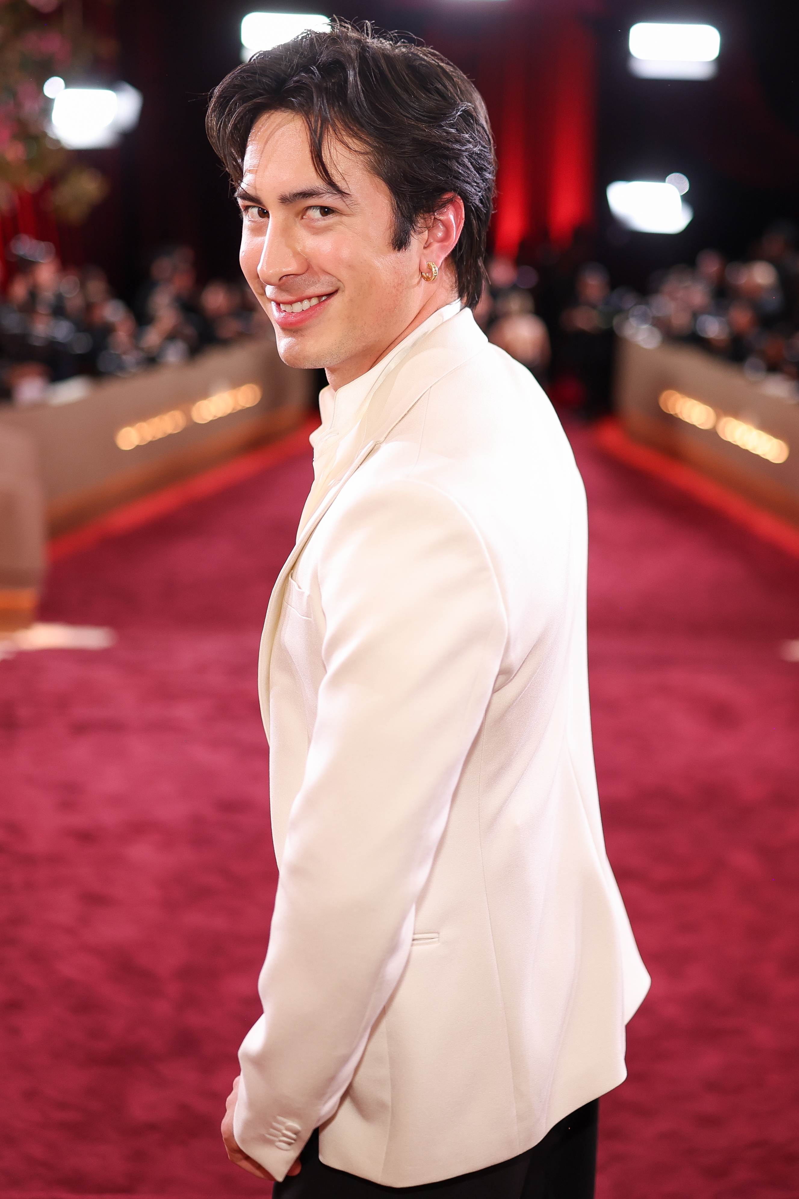 Person i hvit smoking med glatt hår, smiler mens han ser over skulderen på en rød løper-begivenhet