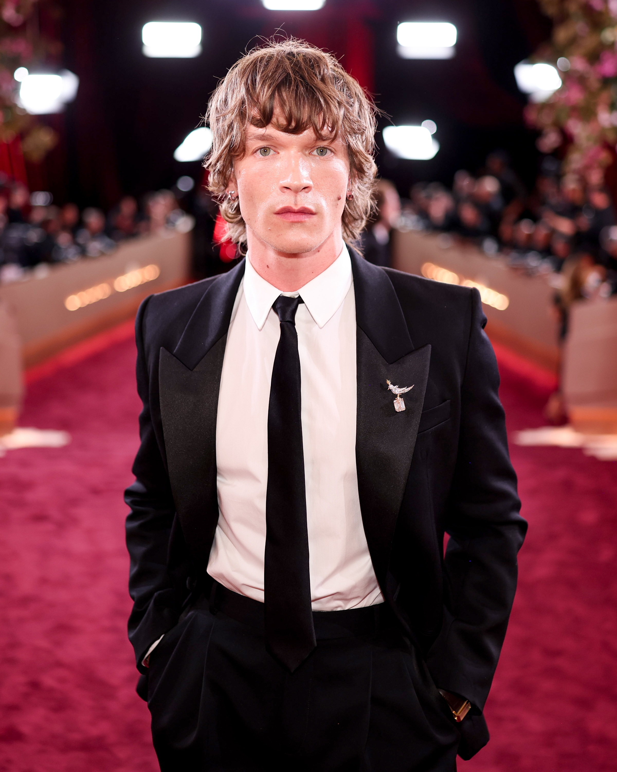 Person i svart dress og slips på en rød løper, iført en brosje, med fotografer og lys i bakgrunnen