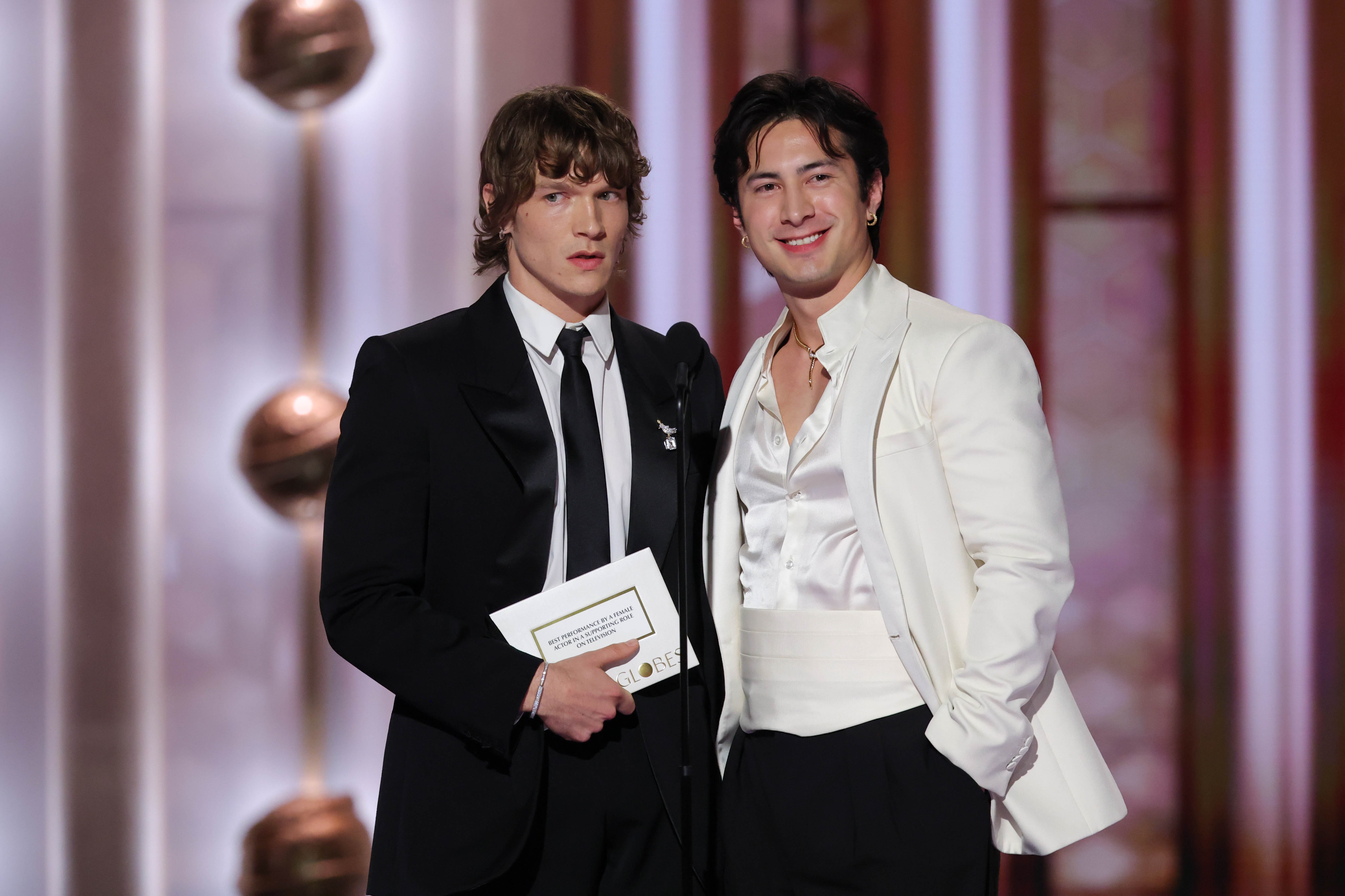 To menn på scenen, den ene i svart smoking holder en konvolutt, den andre i en hvit smokingjakke og skjorte, smilende ved en formell begivenhet