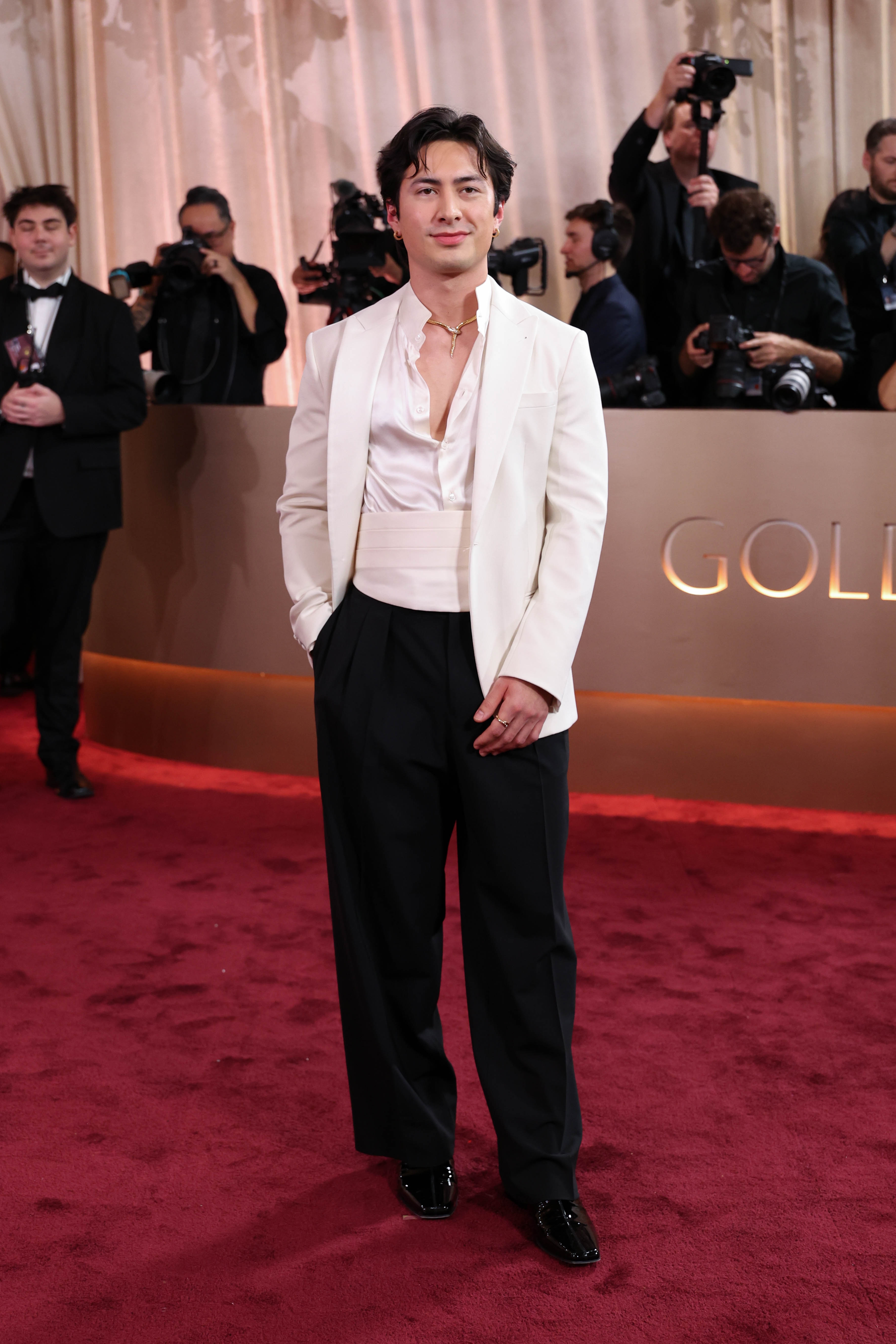 Person i formell antrekk på en rød løper iført en skjorte med åpen krage, hvit blazer, midjebånd og svarte bukser, med fotografer i bakgrunnen