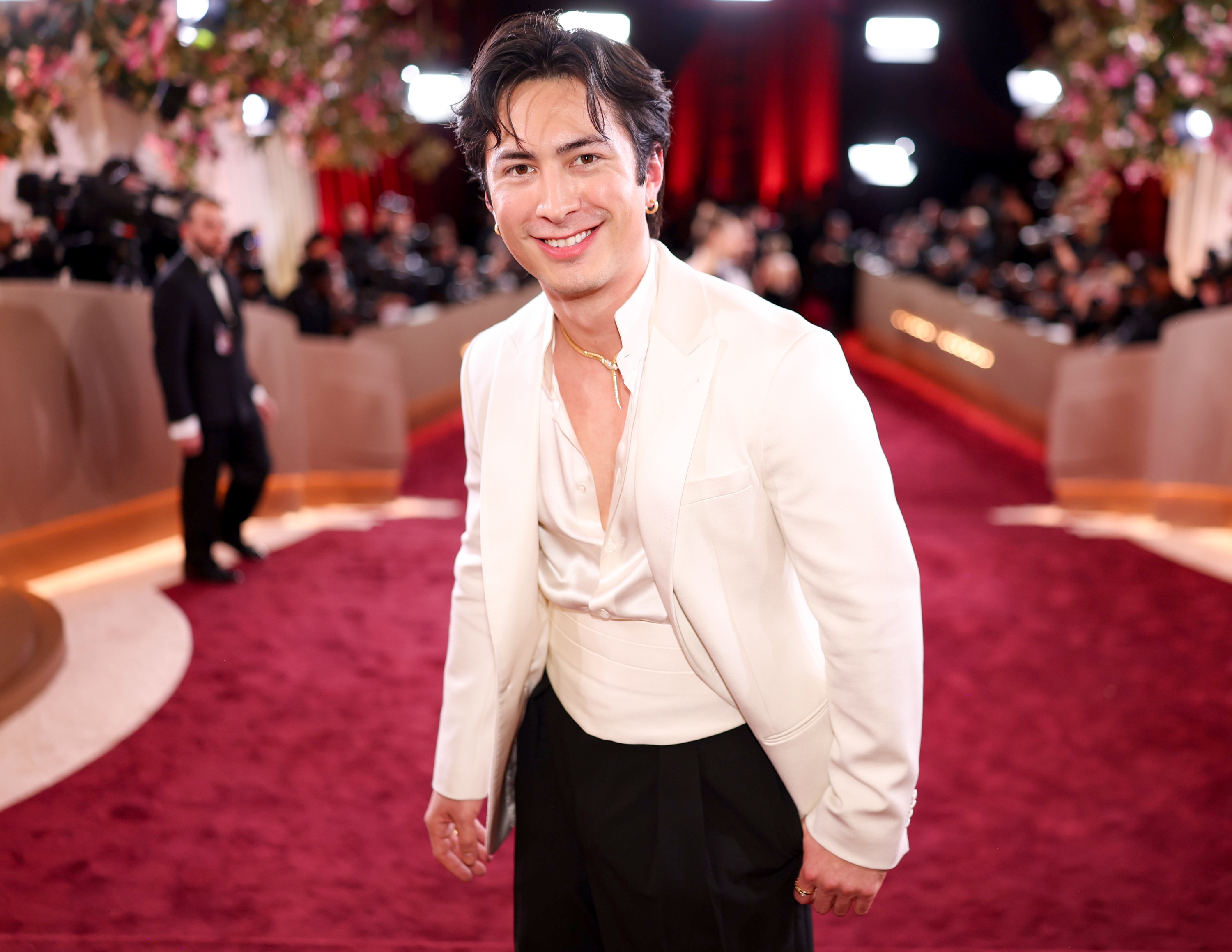 En person i formell antrekk med hvit blazer og elegant skjorte smiler på en rød løper, omgitt av fotografer og elegant innredning