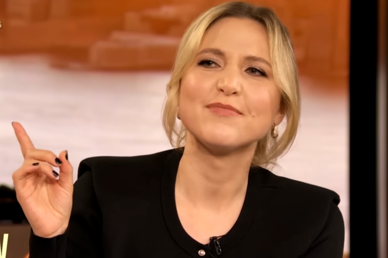 Person smiling with a confident expression, wearing an elegant, sleek outfit. They are gesturing with one hand in a speaking setting