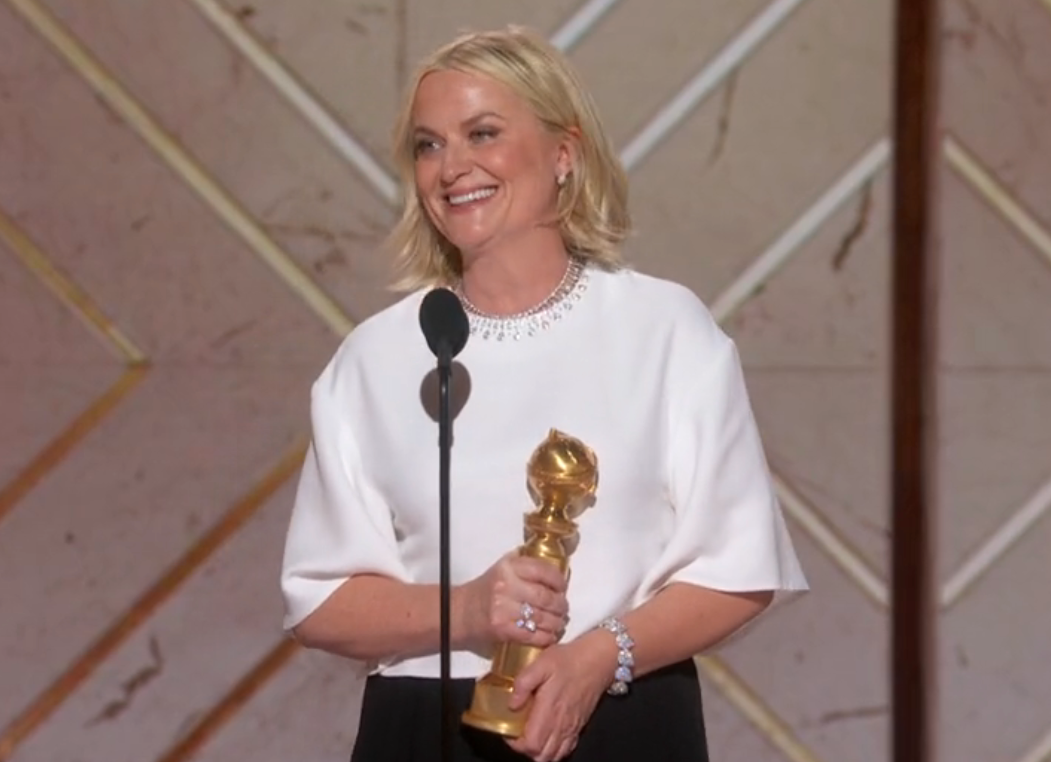 Person successful  a stylish achromatic  blouse holding a aureate  grant  connected  stage, smiling astatine  a microphone during an event